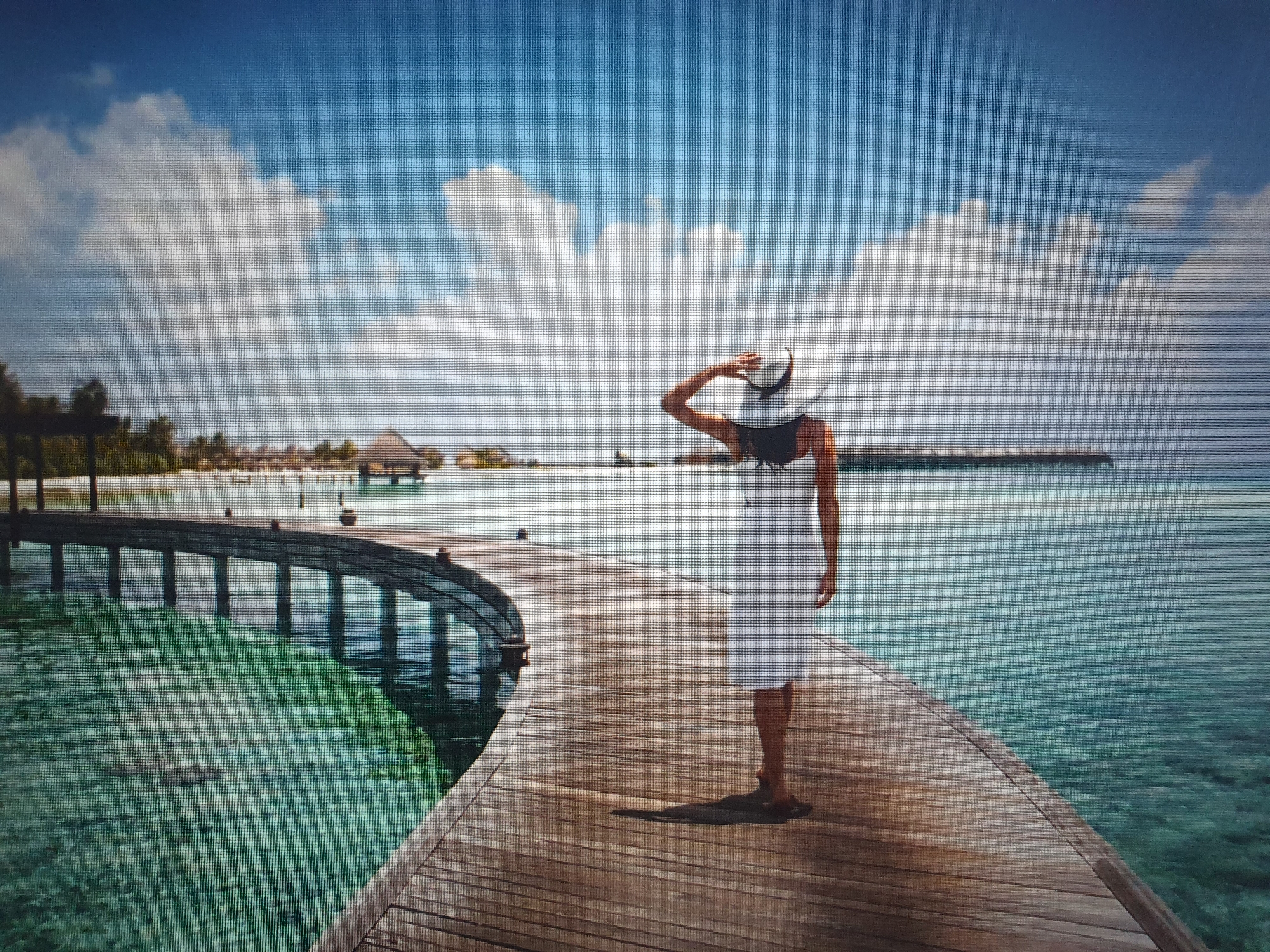 A woman wearing a white dress and a wide-brimmed hat stands on a wooden boardwalk that curves towards the ocean. She is facing away from the camera, with her right hand resting on her hat. The boardwalk is made of weathered wooden planks and appears to extend over turquoise, clear water. In the background, there are overwater bungalows and palm trees. The sky is a vibrant blue with scattered white clouds. The scene depicts a tropical or subtropical island resort. The image does not depict any location in Tashkent, Uzbekistan.