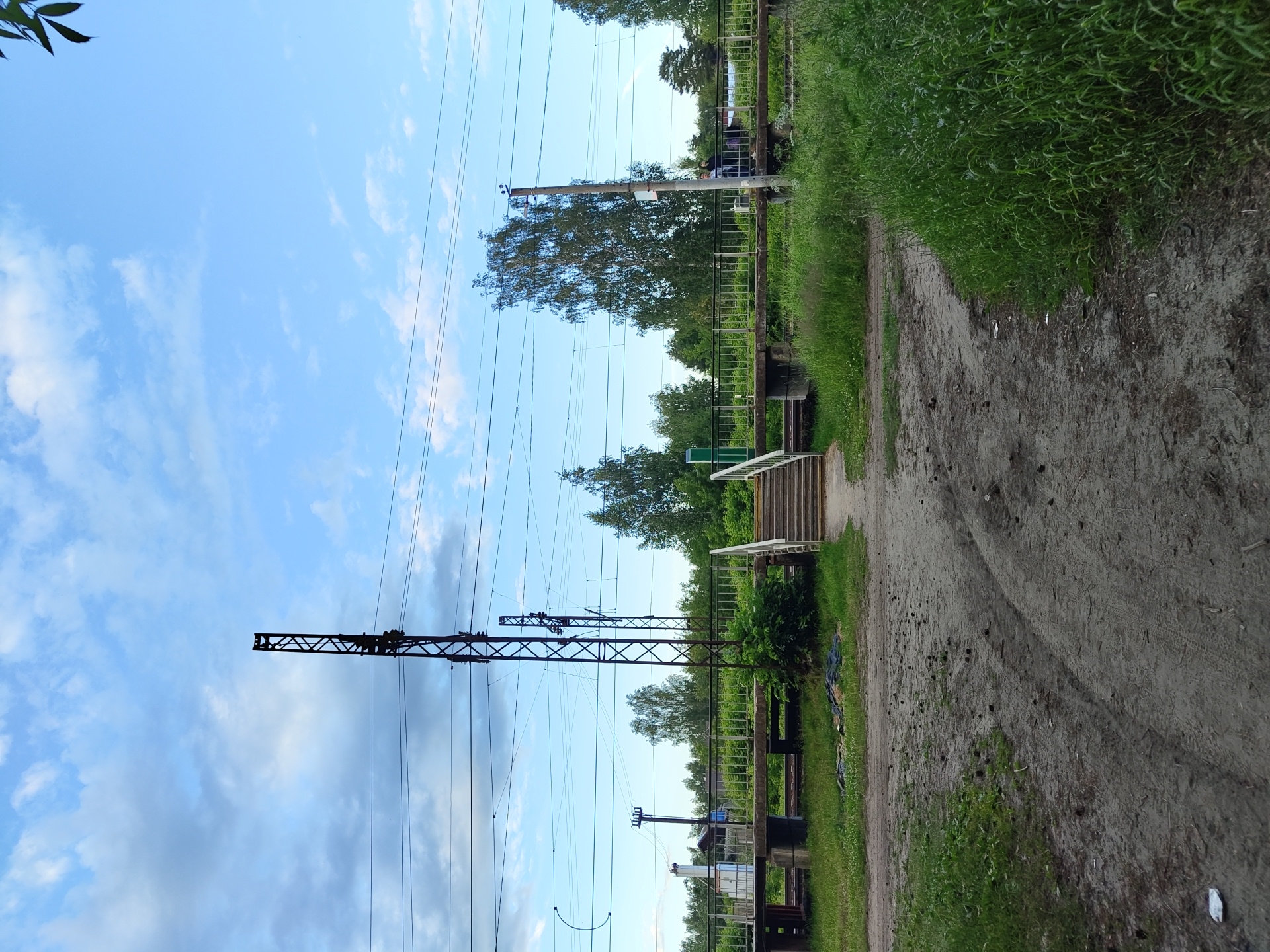 The image depicts an outdoor scene at a railway crossing in Tsibino, Russia. A raised pedestrian overpass with metal railings spans the railway tracks. To the right of the overpass, a set of concrete stairs with metal handrails leads up to it. Two individuals, one appearing to be an adult and the other possibly a child, are standing on the overpass, facing towards the right side of the image. In the foreground, a dirt path with tire tracks curves through overgrown grass. On the left side of the overpass, a white industrial-looking structure is visible behind the railing. Two tall, dark metal electrical pylons with numerous wires are prominent in the scene, one to the left of the overpass and another more centrally located. The sky above is blue with scattered clouds, and a dense line of trees forms the background. The overall impression is of a functional, albeit somewhat rustic, public crossing.