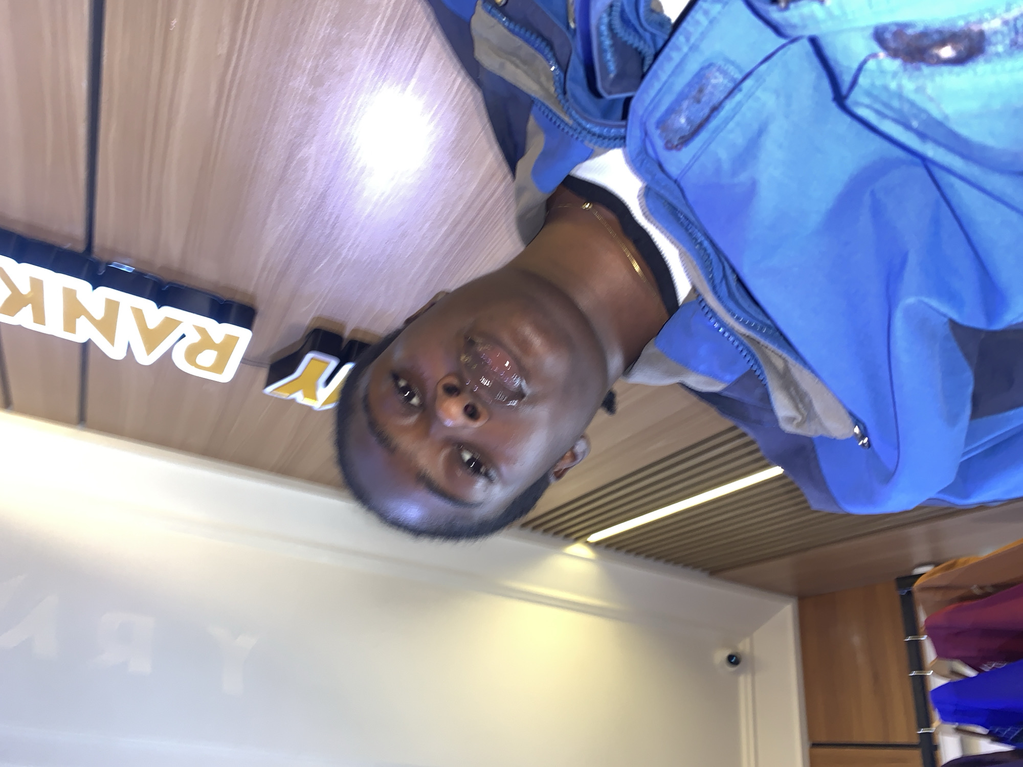 A person is taking a selfie in a store. The person has dark skin and is wearing a blue jacket over a white shirt. The background shows a wooden wall with a lit sign that says "RANK" in yellow letters. There are also clothes hanging on racks in the lower left of the image. The image appears to be taken indoors, possibly in a retail setting. There is no information about the location in Gura Top, Nigeria.