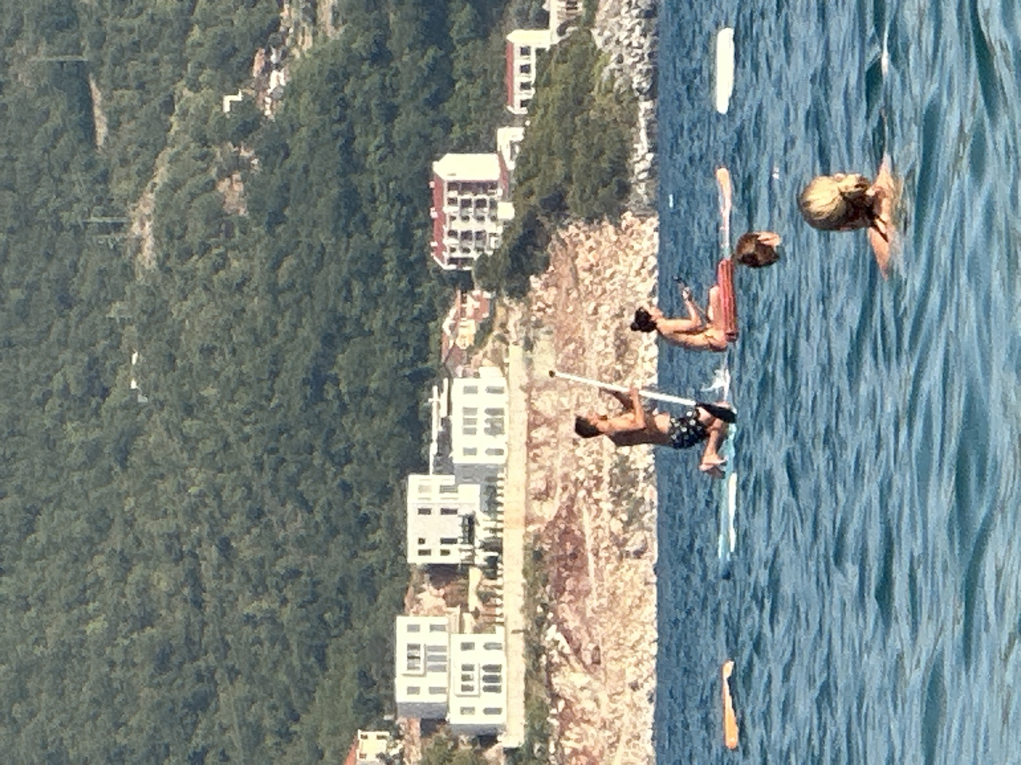 In the city of Boreti, Montenegro, on a clear day, several individuals are seen enjoying water activities. In the foreground, a woman with blonde hair tied back swims in the bright blue, rippling water. Behind her, the head and shoulders of another person are visible, also in the water. Further out, a man is paddleboarding, wearing dark patterned shorts and holding a paddle. To his right, a woman sits on a paddleboard, wearing a swimsuit, and appears to be holding a phone or similar device. Two other paddleboards are visible in the water, one with an orange flotation device. The background features a rocky, sunlit cliff face dotted with greenery, leading up to a hillside covered in dense green trees. Several white modern buildings with multiple stories are situated on the cliffside, overlooking the water. A few more buildings with red-tiled roofs are also visible further up the hillside. The sea is calm with gentle waves.