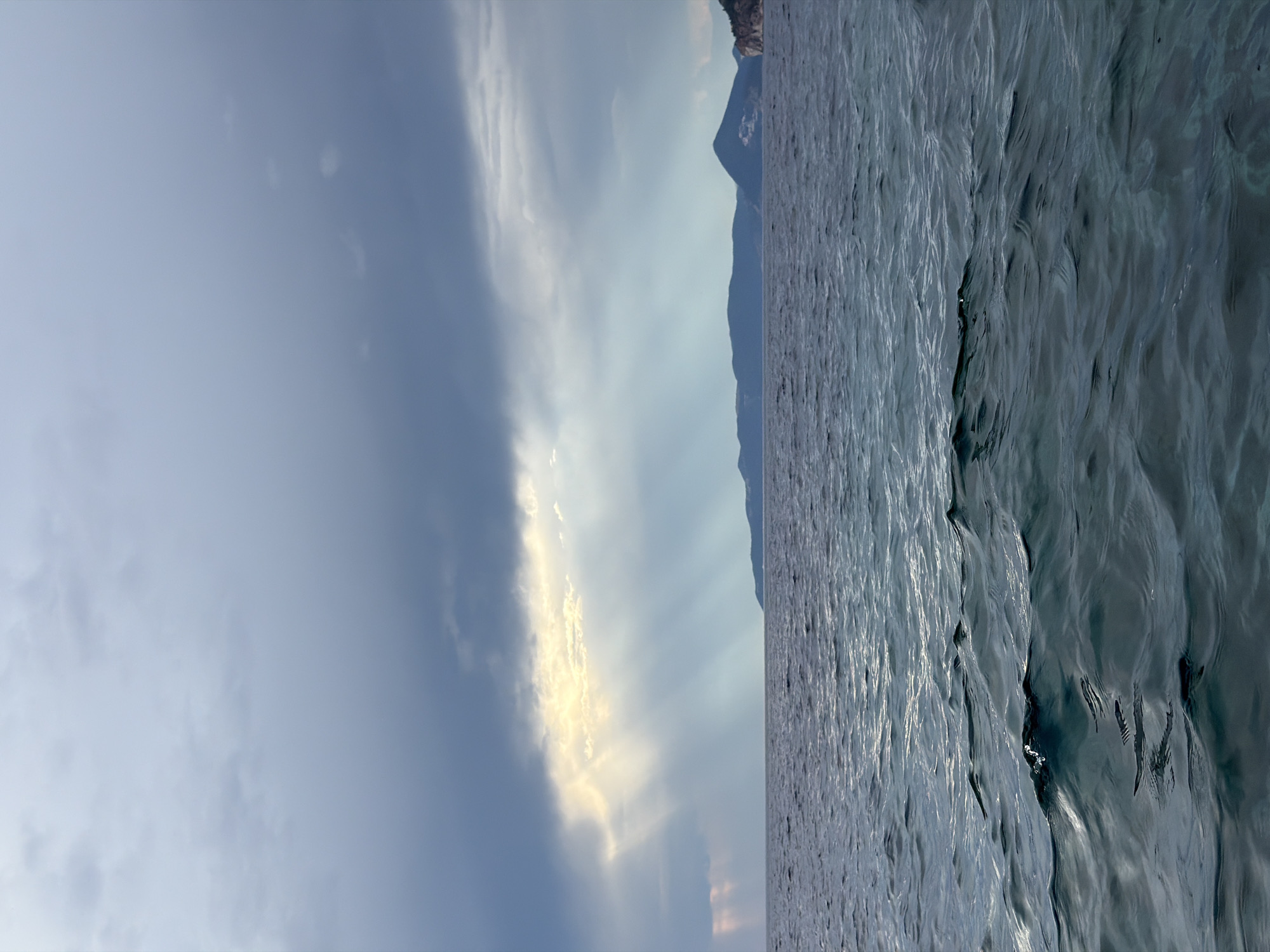 The image captures a serene seascape with a dramatic sky over Petrovac na Moru, Montenegro. In the foreground, the dark blue ocean is agitated by gentle waves, reflecting the light from above. The middle ground features a calm horizon line, separating the water from the land. In the distance, a long, undulating coastline of green hills forms the backdrop. On the far right of the landmass, a few white buildings are visible, suggesting a settlement. The sky is a dominant element, characterized by heavy, dark grey clouds that are partially broken by a bright shaft of golden sunlight. These sunbeams pierce through the clouds, illuminating a section of the sky and casting a warm glow on the distant landscape. The overall impression is one of a fleeting moment of light breaking through an overcast sky, creating a visually striking contrast between light and shadow over the Adriatic Sea.