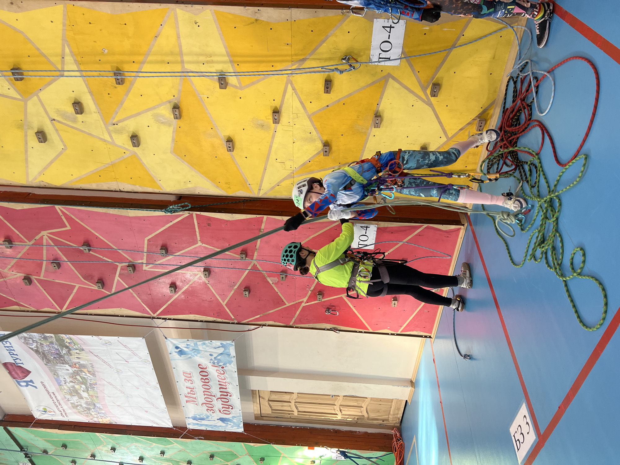 The image shows two people climbing a climbing wall in what appears to be a gymnasium or similar indoor space. The climbing wall is divided into sections of different colors (yellow, red, and green) with geometric patterns. Climbing holds are attached to the wall. A person in a blue climbing suit and helmet is suspended higher up the wall, secured by ropes and a harness. Below them, a person wearing a yellow-green jacket, black leggings, and a helmet is also attached to ropes and a harness, acting as a belayer. There are several ropes coiled on the floor next to the climbing wall. Banners with Russian text and images are hanging on the wall near the climbing wall, with the phrase "Мы за здоровое будущее!" ("We are for a healthy future!"). The location, Berezniki, Russia, is implied from the OCR text "Туризм" and the banners. "TO-4" is written on two rectangular white signs attached to the yellow section of the wall. The floor is blue with red lines marking boundaries. The text "63 3" is written in white on a white square in the lower right-hand corner of the image.