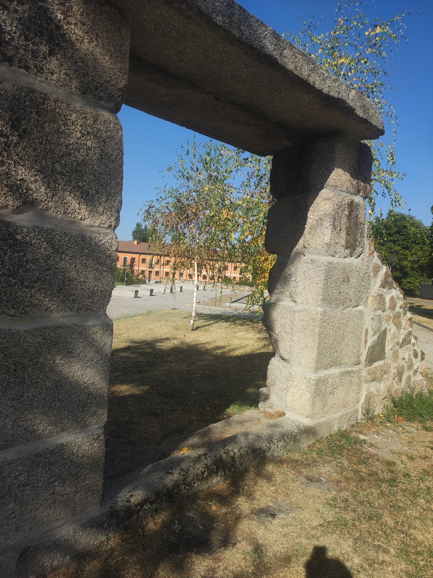 The image shows a close-up view of a rough-hewn stone archway, framing a scene with a building and trees in the background. The archway is constructed from large, irregular granite blocks, with some smaller stones used in its construction. A light-colored mortar is visible between the stones. Through the archway, a long, orange building with multiple windows is visible, set against a clear blue sky. In front of the building is a paved area with a few stone benches. A slender birch tree with yellowing leaves stands in the foreground, to the right of the archway. The ground in front of the archway is covered in dry grass and fallen leaves. The overall impression is one of an outdoor setting, possibly in a park or historical site, given the architectural style of the archway and the presence of the building. The location is identified as Real Sitio de San Ildefonso, Spain. The image is captured in daylight, with shadows indicating the sun's position. There is no indication of any people actively engaged in the scene, though a shadow of a person is visible in the lower right corner of the frame, suggesting someone is taking the photograph.
