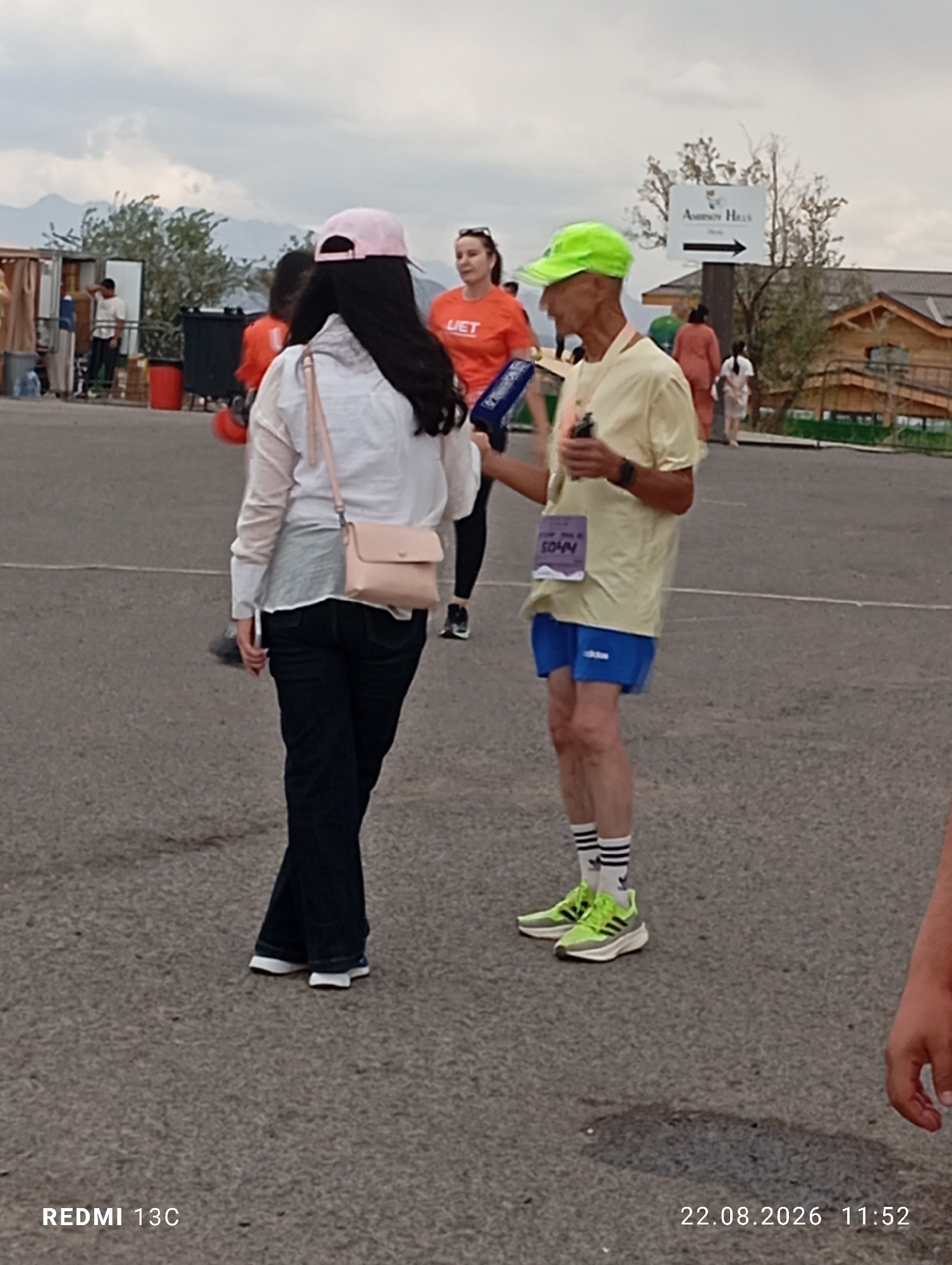 The image depicts an outdoor scene at the Amirsay ski resort in Uzbekistan. In the foreground, a man wearing a lime green cap, a yellow t-shirt, blue shorts, and striped socks stands on asphalt. He is holding a blue object, possibly a communication device or a microphone, and has a race bib with the number "5044" attached to his shirt. To his left, a woman wearing a white shirt, dark pants, and a pink cap is seen from the back. She is carrying a light pink crossbody bag. In the background, a directional sign points to "AMIRSAY HILLS" with an arrow. Several other individuals and structures, including a wooden building and tents, are visible in the distance. The overall atmosphere suggests an event or gathering taking place at the resort.