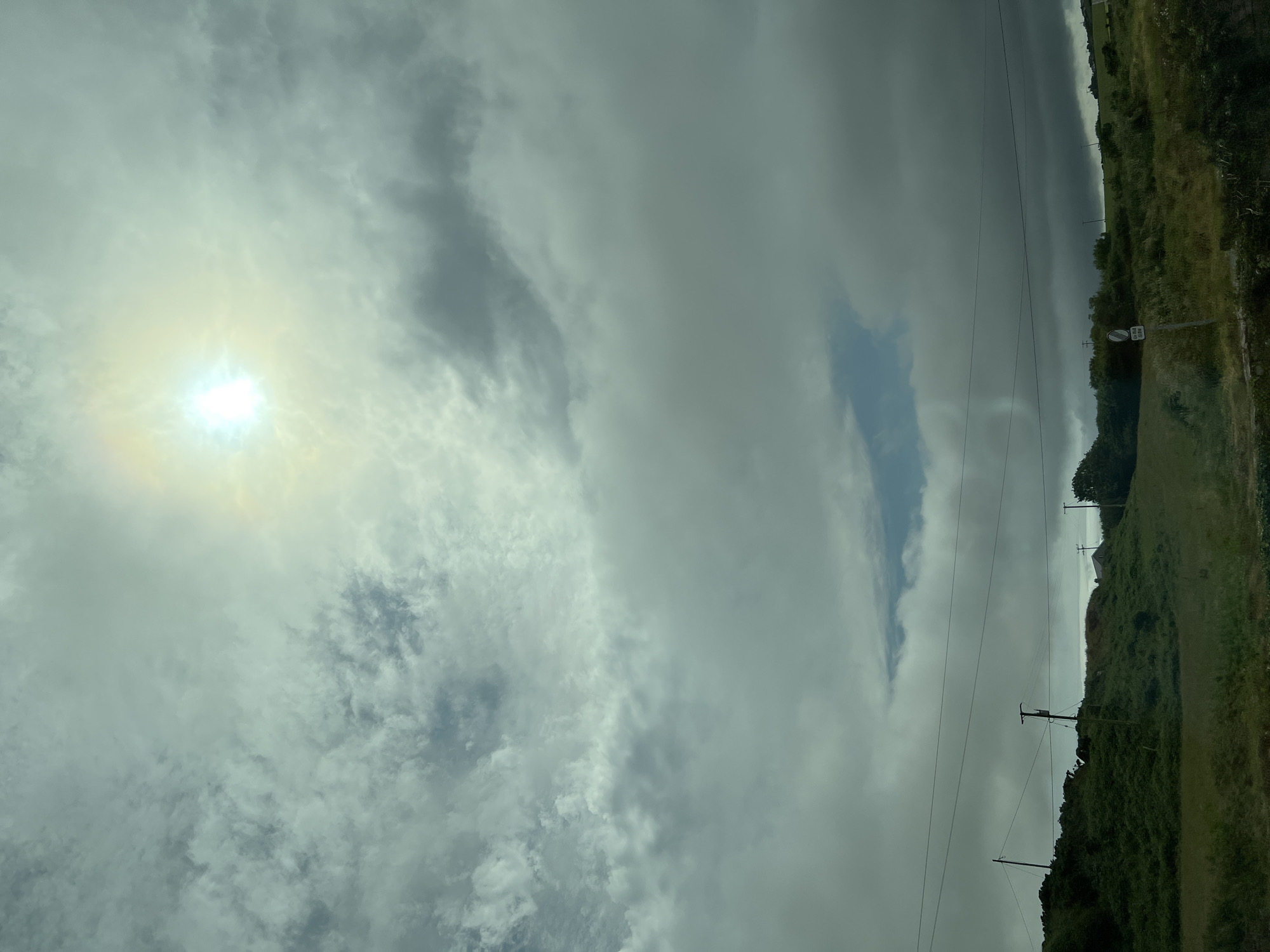 This media file captures an overcast sky with a bright, hazy sun partially obscured by clouds, casting a soft, diffused light. Wispy, grey clouds dominate the sky, with a patch of clearer blue sky visible in the upper right portion.
The scene transitions to a green, hilly landscape on the right side of the image. The terrain is covered with lush grass and dense foliage, with scattered trees dotting the hills. A few power lines stretch across the frame, supported by wooden poles. In the distance, along the horizon, is a faint indication of a building or structure. A white circular sign with black text and a red border is visible on a pole in the mid-ground.
The overall impression is of a slightly moody, perhaps humid, day in a rural, coastal environment. The time of day appears to be either morning or late afternoon, given the angle and quality of the sunlight. The landscape suggests a location with rolling hills and an expansive sky, consistent with the provided location context of Dunfanaghy, Ireland. There are no people or obvious animals visible in the frame. The activity taking place, if any, is not evident, as the image appears to be a still landscape shot, possibly taken from a moving vehicle.
