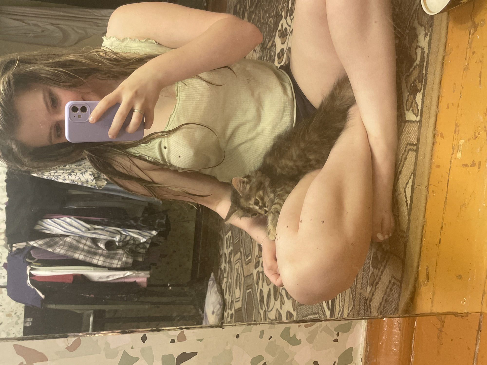 A young woman is taking a selfie in a dimly lit room with a mirror reflecting her and a small kitten. She is sitting cross-legged on a patterned rug, holding a light green tank top. The kitten is nestled in her lap. The background shows a clothes rack with several items of clothing and a cluttered shelf. The floor is made of distressed wooden planks. The scene appears to be indoors, possibly a bedroom or a small apartment.