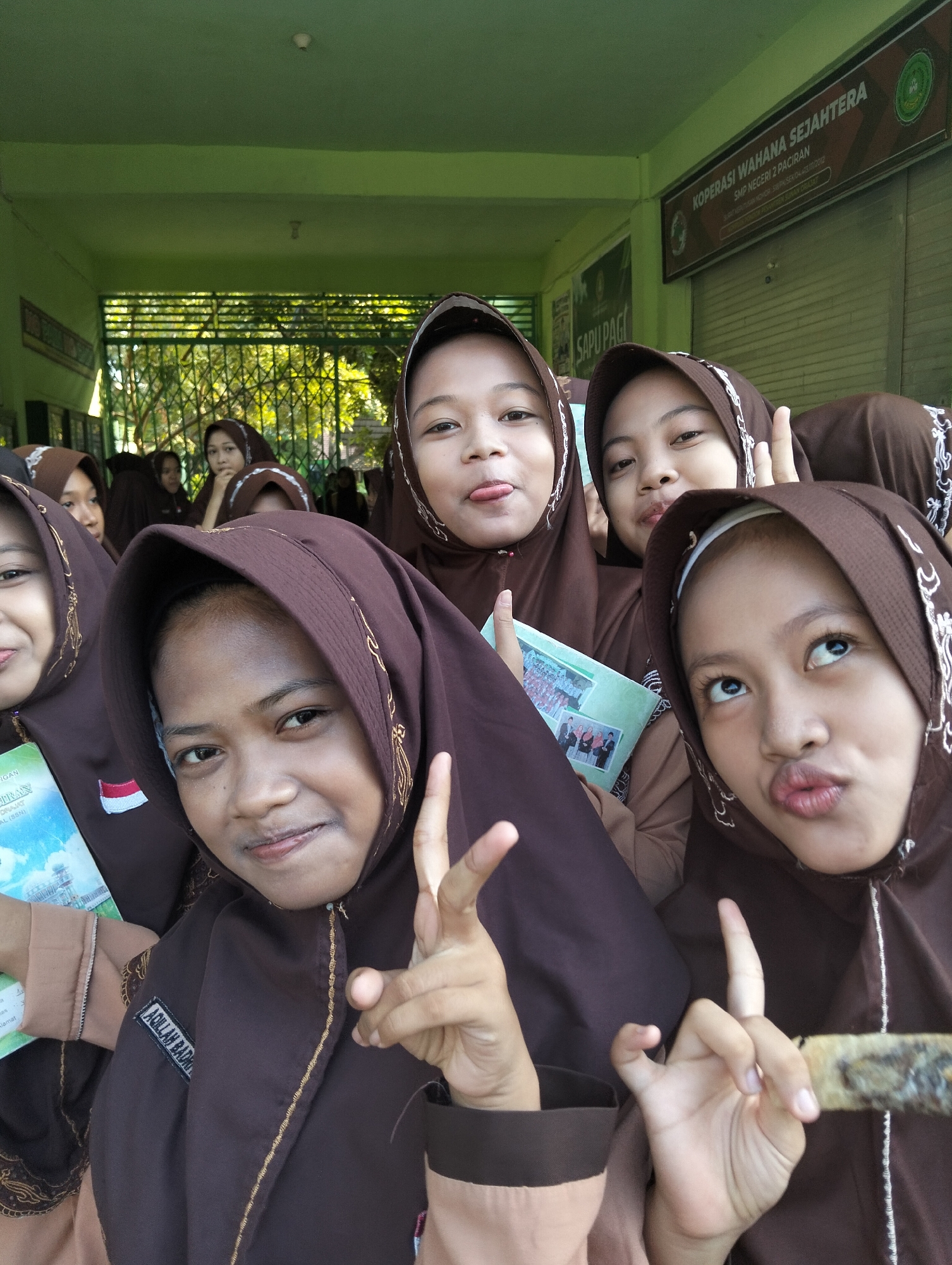 A group of young female students, likely in their early teens, pose for a selfie in front of a green building. They are all wearing brown hijabs and uniforms. The students are making playful facial expressions, with some sticking out their tongues and others puckering their lips. In the background, a sign reads "KOPERASI WAHANA SEJAHTERA SMP NEGERI 2 PACIRAN," indicating the location as Paciran, East Java, Indonesia. The students appear to be on school grounds, possibly during a break or event. One student on the left holds a booklet or folder with a picture of a group of people on it. The overall scene suggests a lighthearted and youthful moment captured outdoors.