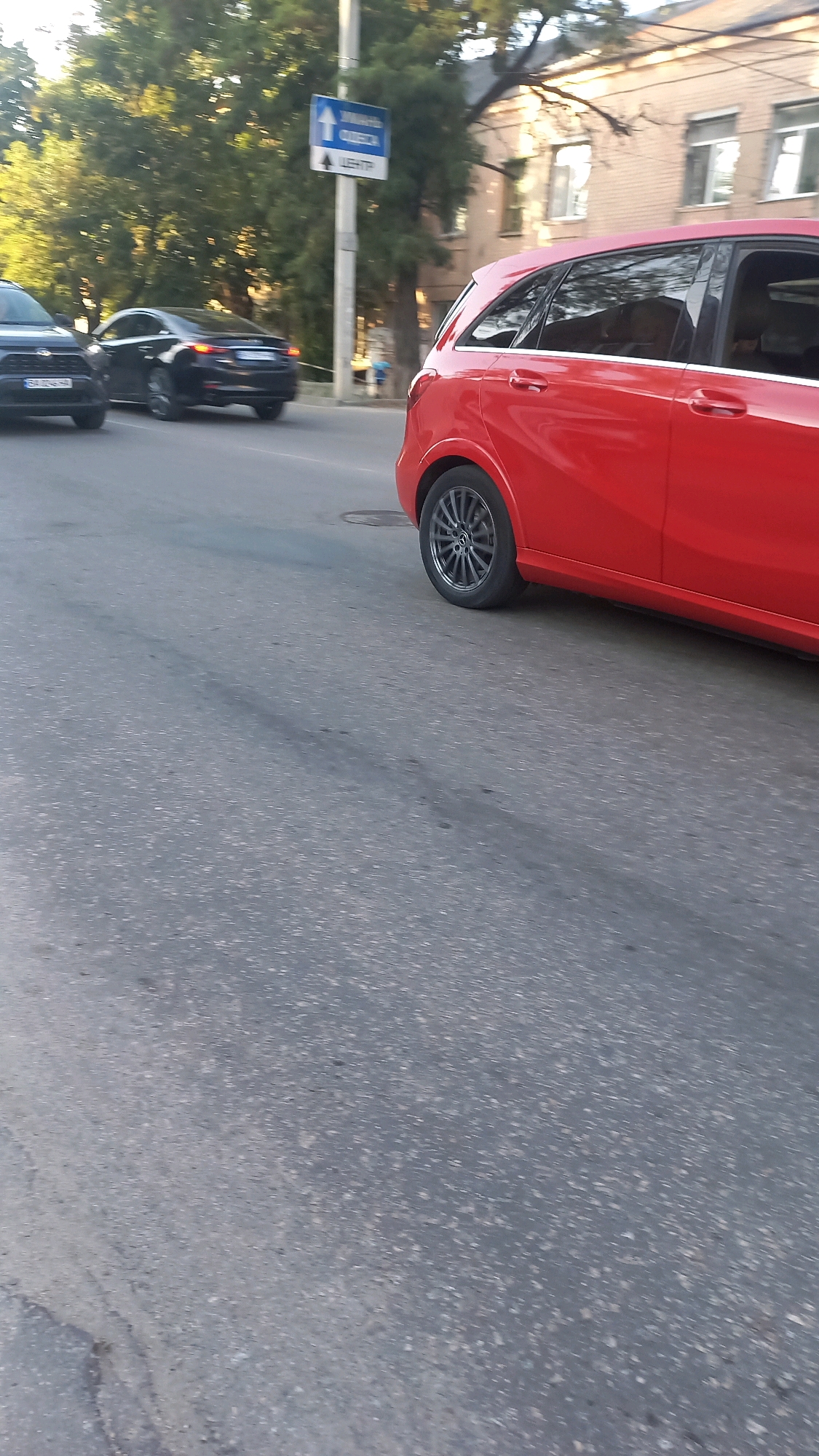 The image shows a street scene in Kropyvnytskyi, Ukraine. A red hatchback car is in the foreground, traveling on asphalt. Behind it, a black sedan is also on the road. A white signpost with blue directional arrows and Ukrainian text, indicating a route to "CENTER," is visible on the left side of the road. Trees and buildings form the background. The lighting suggests it is daytime.