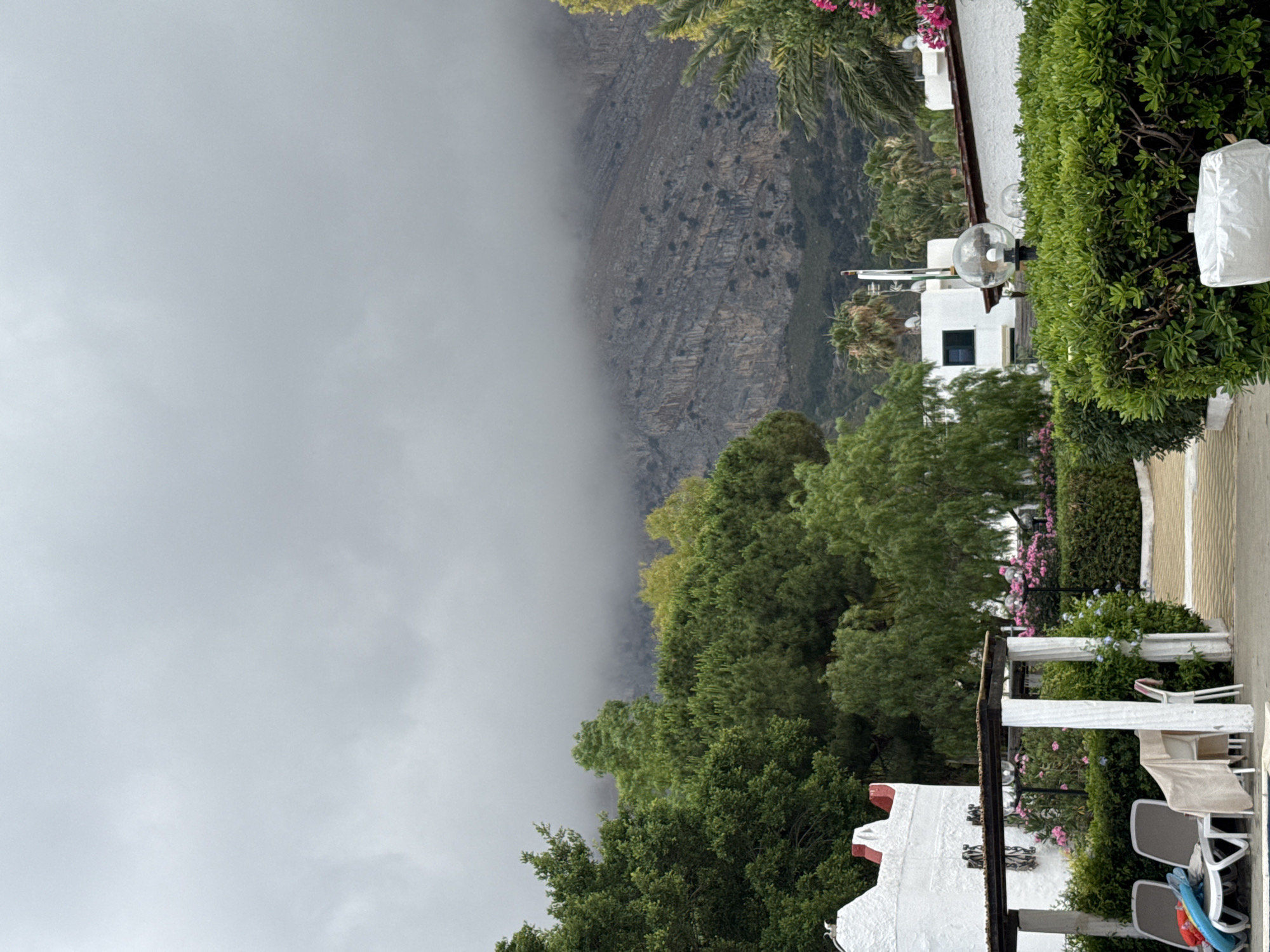 The image shows an outdoor scene with a cloudy sky. In the foreground, there are white lounge chairs by a pool, with towels draped over them. There's a white pergola with white pillars and a wooden roof. Beyond this, a white building with red accents is visible. Lush green trees and bushes are abundant, with some pink flowers interspersed. In the mid-ground, more white buildings with brown roofs and palm trees are present. In the background, a large, rocky mountain is shrouded in mist and clouds. The scene appears to be a resort or hotel complex in Isola delle Femmine, Italy, given the architecture and vegetation, and the presence of a pool suggests a vacation setting. There are no people visible in the image.