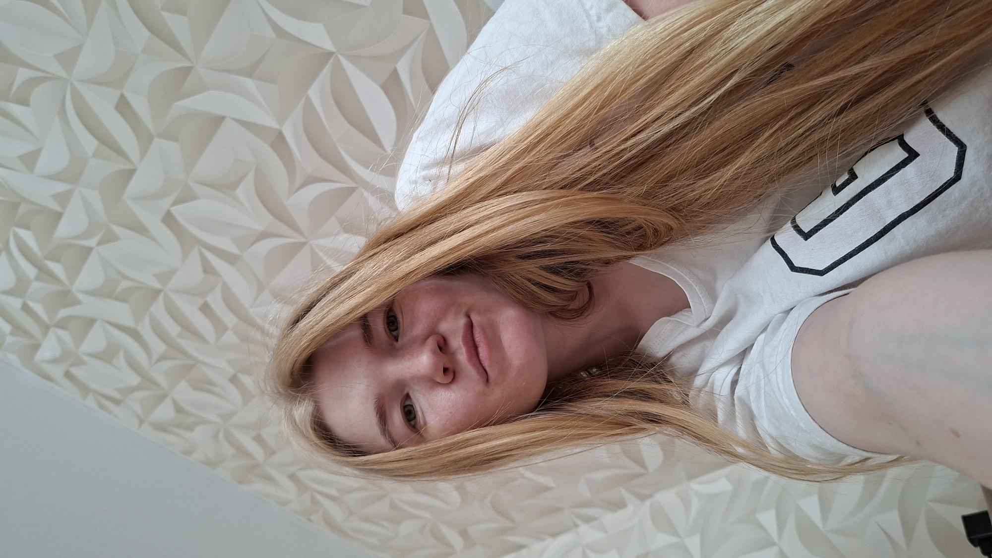 The media file is a portrait of a young woman with long, blonde hair. She is lying down, possibly in bed, as indicated by the pillow and the overall relaxed pose. Her head is tilted, and her hair is spread out around her. She is wearing a white t-shirt with a black graphic design on the sleeve, which appears to be a stylized letter or symbol. The background is a textured wall with a geometric pattern in light beige and white tones. The lighting suggests it is daytime, but no specific time or weather conditions are discernible. The woman has a gentle smile, and her expression conveys a sense of calm and contentment. The image is taken from a slightly elevated angle, looking down at the subject. No specific location cues are visible other than the geometric pattern on the wall. There is no visible text in the image.