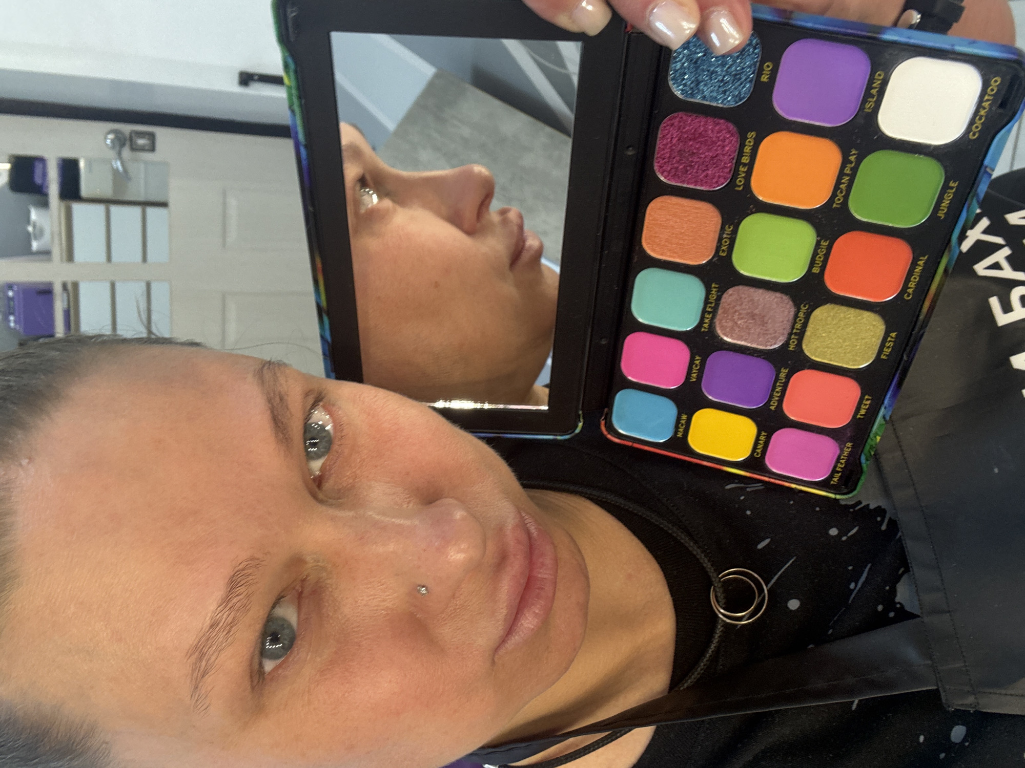 The image is a close-up selfie taken by a person holding a makeup palette. The palette has a variety of bright colors and is labeled with names such as "RIO," "ISLAND," "COCKATOO," "JUNGLE," "CARDINAL," "FIESTA," "TWEET," "TAIL FEATHER," "CANARY," "ADVENTURE," "MACAW," "VAYCAY," "TAKE FLIGHT," "EXOTIC," "LOVE BIRDS," "BUDGIE," "TOCAN PLAY," and "HOT TROPIC." A mirror is attached to the palette, reflecting the person's face in profile. The person has fair skin, light eyes, and a piercing on the side of their nose. They are wearing a dark-colored top with a splatter design and a necklace with a ring pendant. The background shows a portion of a white wall with paneling and a mirror. The text 'БАЛ' is visible on a black surface near the bottom right corner of the image. There is no evidence to suggest the location is in Perm, Russia.
