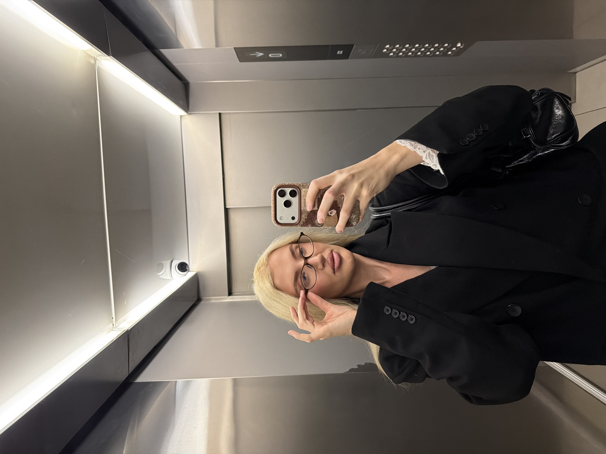 An indoor scene depicts a blonde female taking a mirror selfie inside an elevator, located in Belgrade, Serbia. The elevator cab features brushed metallic gray walls and ceiling, illuminated by linear LED strip lighting along the ceiling edges. A white dome-shaped CCTV camera is mounted in the upper left corner of the cab. On the right wall, a dark control panel with an illuminated digital display shows "0" and a downward arrow, indicating the elevator's current floor or direction. Below the display are rows of circular call buttons.
The female subject, positioned centrally, is holding a smartphone with a distinctive patterned brown case and a triple-lens camera system in her right hand, pointed towards her reflection to capture the image. Her left hand is raised, adjusting or holding a pair of oval, dark-rimmed eyeglasses at the bridge of her nose. She wears a black double-breasted blazer, and a white lace cuff is visible on her right wrist underneath the blazer sleeve. A small, black textured bag is slung over her right forearm or shoulder. Her blonde hair is long and falls over her shoulders.
