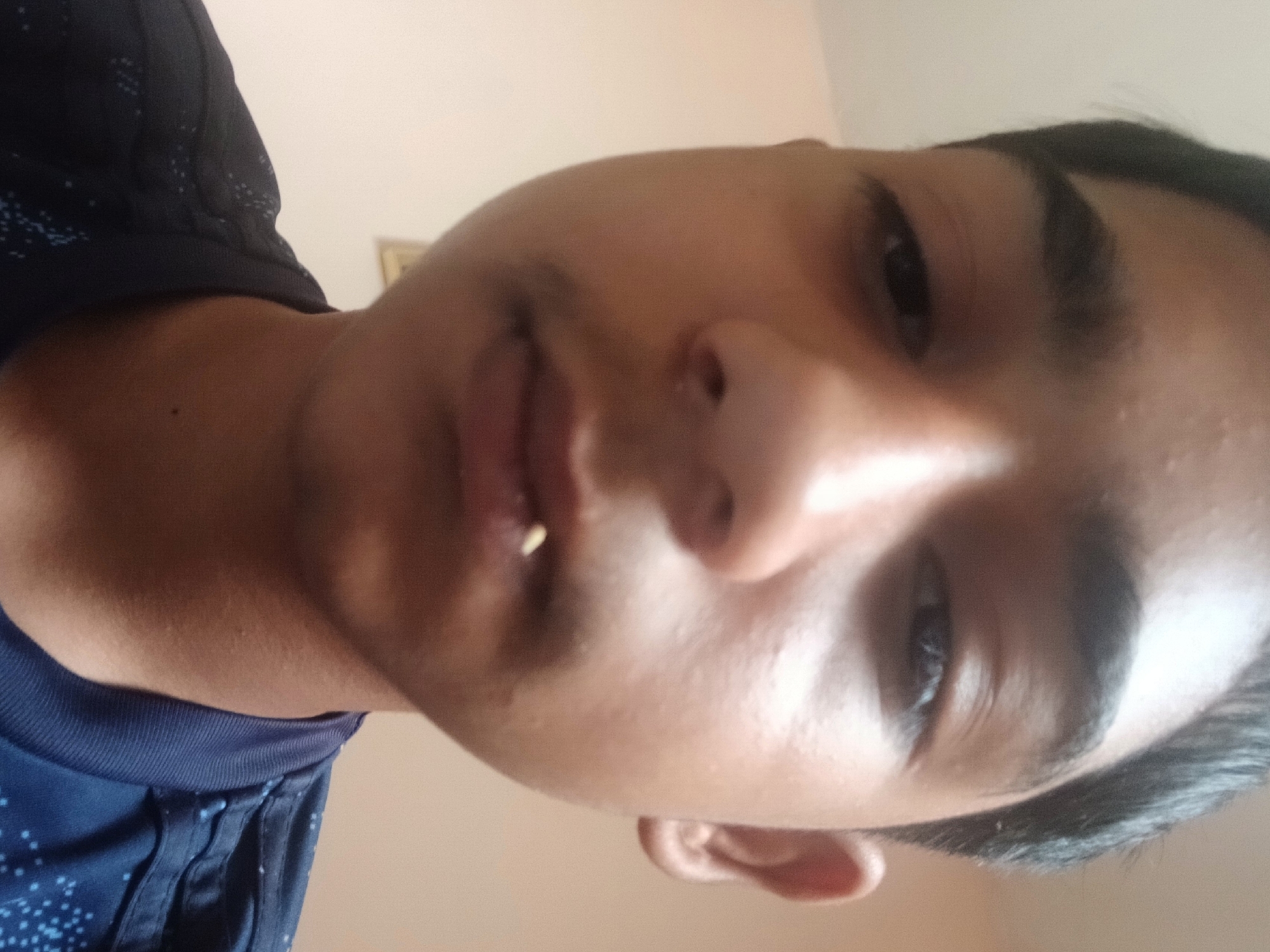 The image shows a close-up selfie of a young male individual. The camera angle is low, looking upwards toward the subject's face. The individual has dark hair, dark eyes, and a prominent nose. Part of a blue t-shirt with a dotted pattern is visible in the lower left corner of the frame. A small white object is visible between the lips. The background is a plain, light-colored wall. There is no information available to determine the location is in Karanovo, Bulgaria.