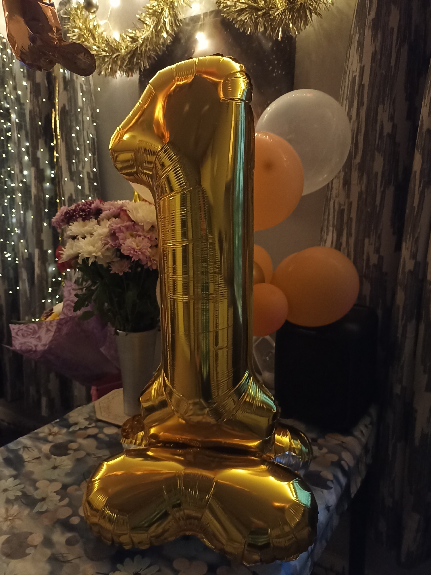 A gold foil balloon shaped like the number "1" is the central subject of the image. It stands upright on a table covered with a floral-patterned tablecloth. Behind the balloon, a bouquet of white and pink flowers in a silver vase is visible. In the background, there are several balloons: a large white one, a pale orange one, and two smaller orange ones. A black object, possibly a speaker or box, is partially obscured by the balloons. A string of white fairy lights is draped across the top of the image, and a gold tinsel garland is also present. The scene appears to be indoors, possibly set up for a celebration. There is no information available that places this image in Annagry, Ireland.