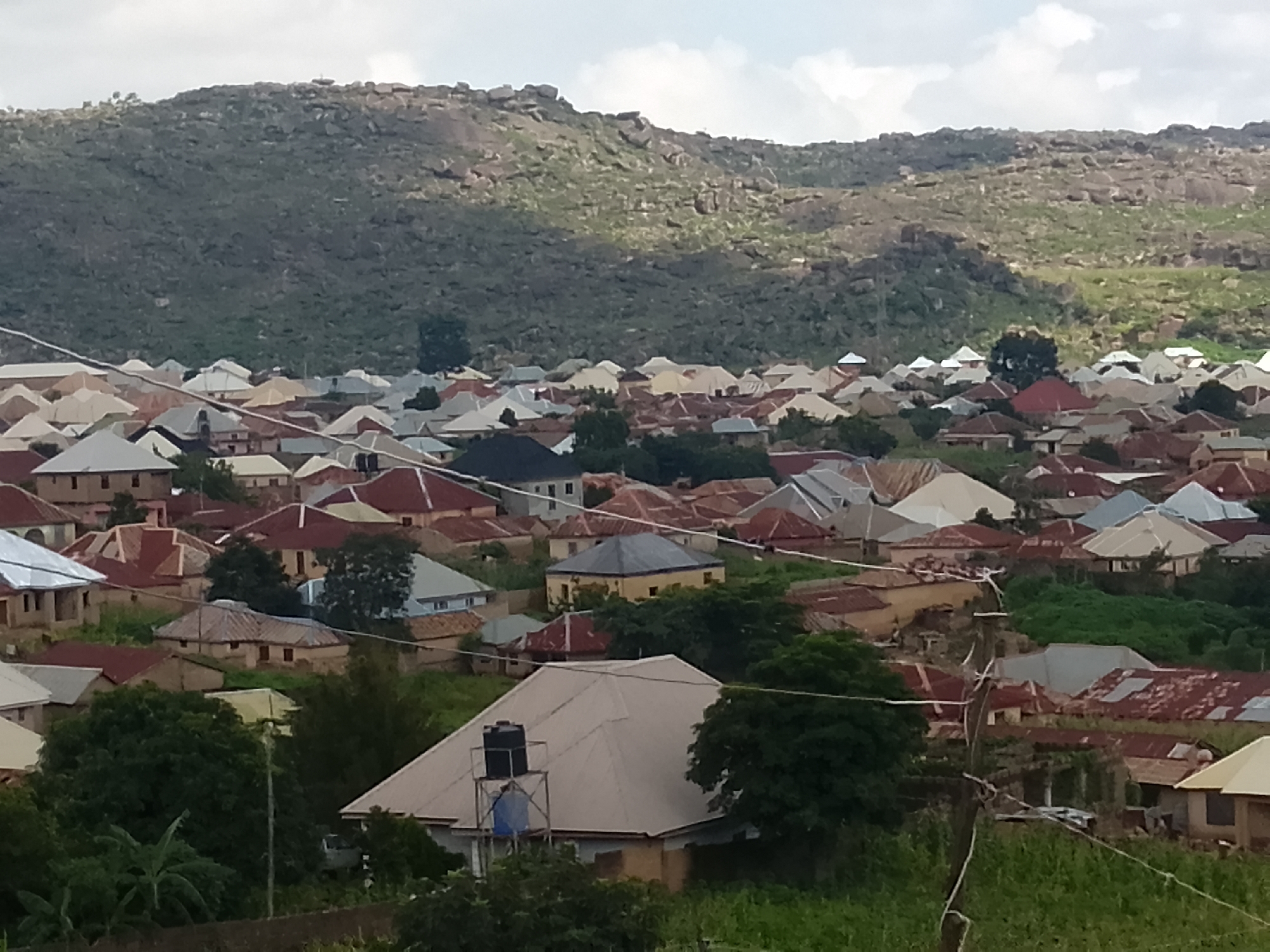The image shows a dense residential area in Rukuba Road, Nigeria, nestled at the foot of a rocky, vegetated hillside. The houses are primarily single-story structures with corrugated metal roofs of varying colors, including red, silver, and beige. Many of the roofs feature distinct triangular or pyramidal shapes. The buildings are clustered closely together, with narrow pathways or unpaved areas between them. Vegetation, including trees and shrubs, is interspersed throughout the settlement, particularly on the slopes of the hill and around some of the houses. A water tank is visible on a stand on one of the rooftops in the foreground. The sky is overcast with scattered clouds. The scene depicts a typical urban or semi-urban landscape in the region, characterized by its built environment and natural surroundings.