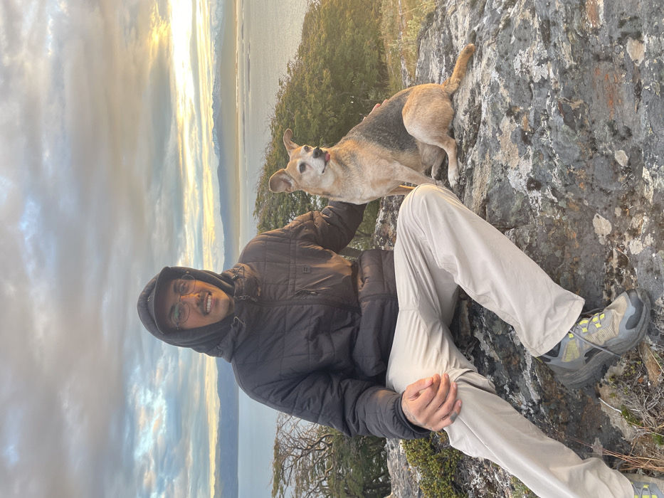 The image shows a man and a dog posing for a photo on a rocky hillside overlooking a body of water and distant mountains. The original image is rotated 90 degrees to the left, depicting the scene sideways.
In the foreground, a man is seated or crouched on a dark, textured rock surface. He is wearing a dark, possibly quilted, hooded jacket and light-colored hiking pants. He has glasses and a warm smile on his face, looking directly at the camera. His right hand is resting on his leg. His feet are visible, shod in robust hiking shoes with grey and bright green accents.
Next to the man, a medium-sized dog stands on the rocky ground, also facing the camera. The dog has light brown fur with darker markings on its back and around its face. Its ears are pointed and erect, and its tongue is slightly out, giving it an engaged or happy expression. The man's left arm is close to the dog, suggesting he might be petting it or just having it sit beside him.
The background reveals a expansive natural landscape. Beyond the immediate rocky and sparsely vegetated terrain, there's a wide body of water, likely a lake or ocean, stretching towards the horizon. Across the water, distant mountains or landmasses are visible, appearing somewhat hazy. The sky above is cloudy, with patches of bright, warm light breaking through, suggesting either sunrise or sunset, casting a soft, golden glow on the clouds and reflecting slightly on the water. The overall atmosphere is serene and natural, indicating an outdoor adventure or leisurely stop. There is no visible text in the image.