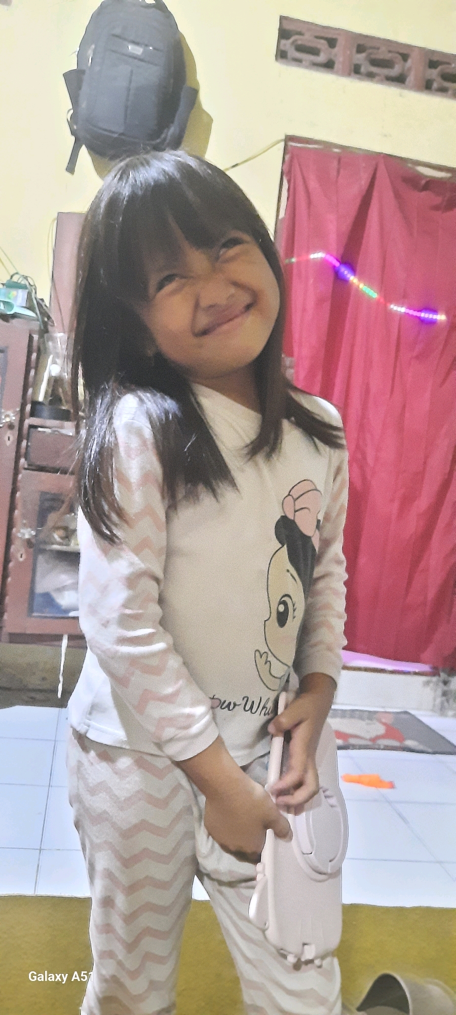 A young girl wearing pajamas with a chevron pattern and a white long-sleeved shirt featuring a cartoon character is standing in what appears to be a residential interior. She is holding a light pink object in her hands, which resembles a small electronic device or a toy. The background includes a red curtain, a backpack hanging on the wall, a cabinet, and tiled flooring. A watermark in the lower-left corner indicates the image was taken with a "Galaxy A51." The scene suggests an informal indoor setting in Jatitengah, Indonesia.