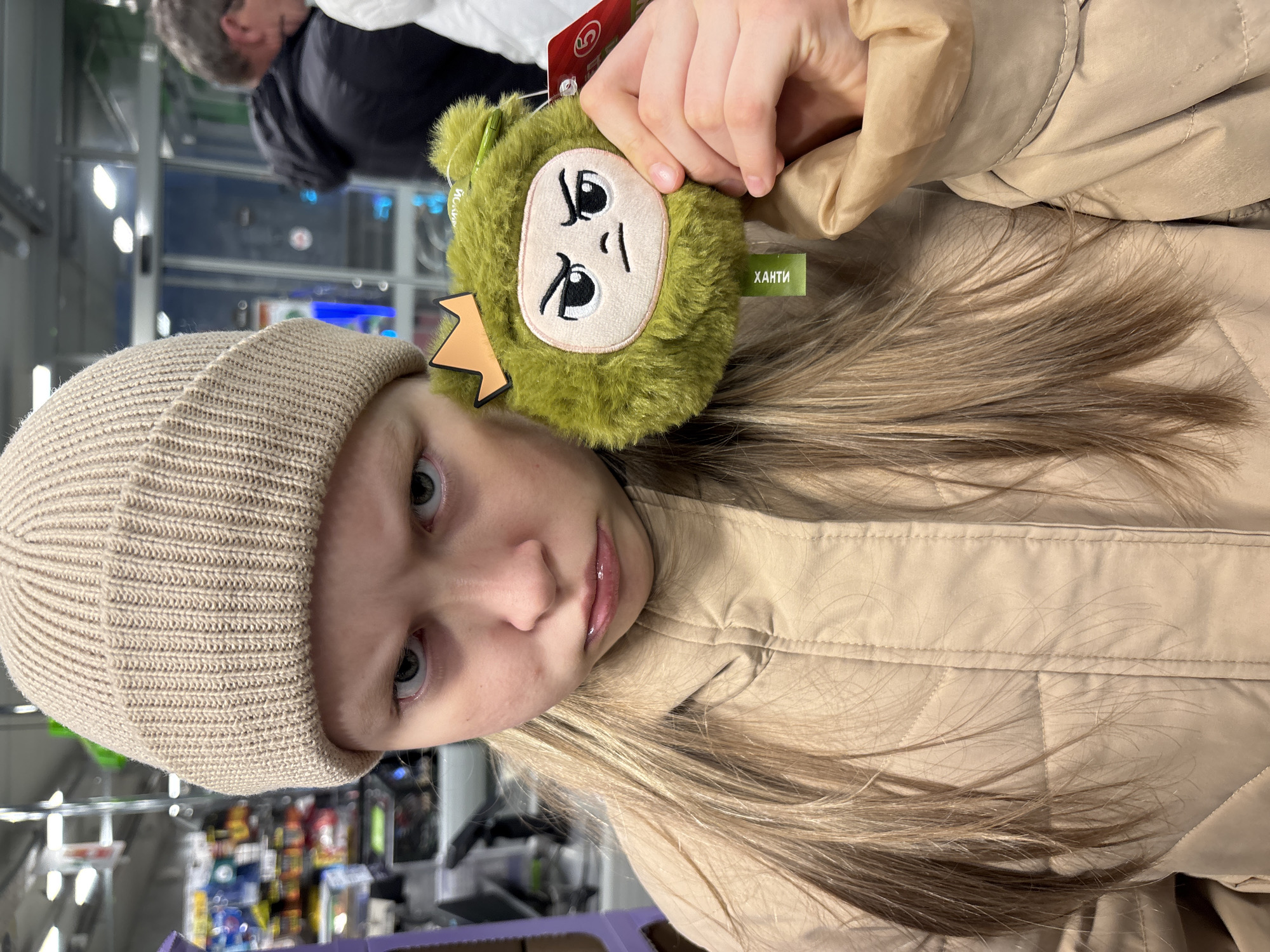 A fair-skinned young person with light blonde hair and gray-green eyes, wearing a beige knit cap and a quilted beige jacket, is holding up a small, green, plush toy with a frowning face and a tag labeled "ХАНТИ". The background includes commercial shelves with various products and a blurred figure of a person with glasses. Based on the label "ХАНТИ", the location is suggested to be in the city of Ягул, Russia, given that the language on the tag is Russian.
