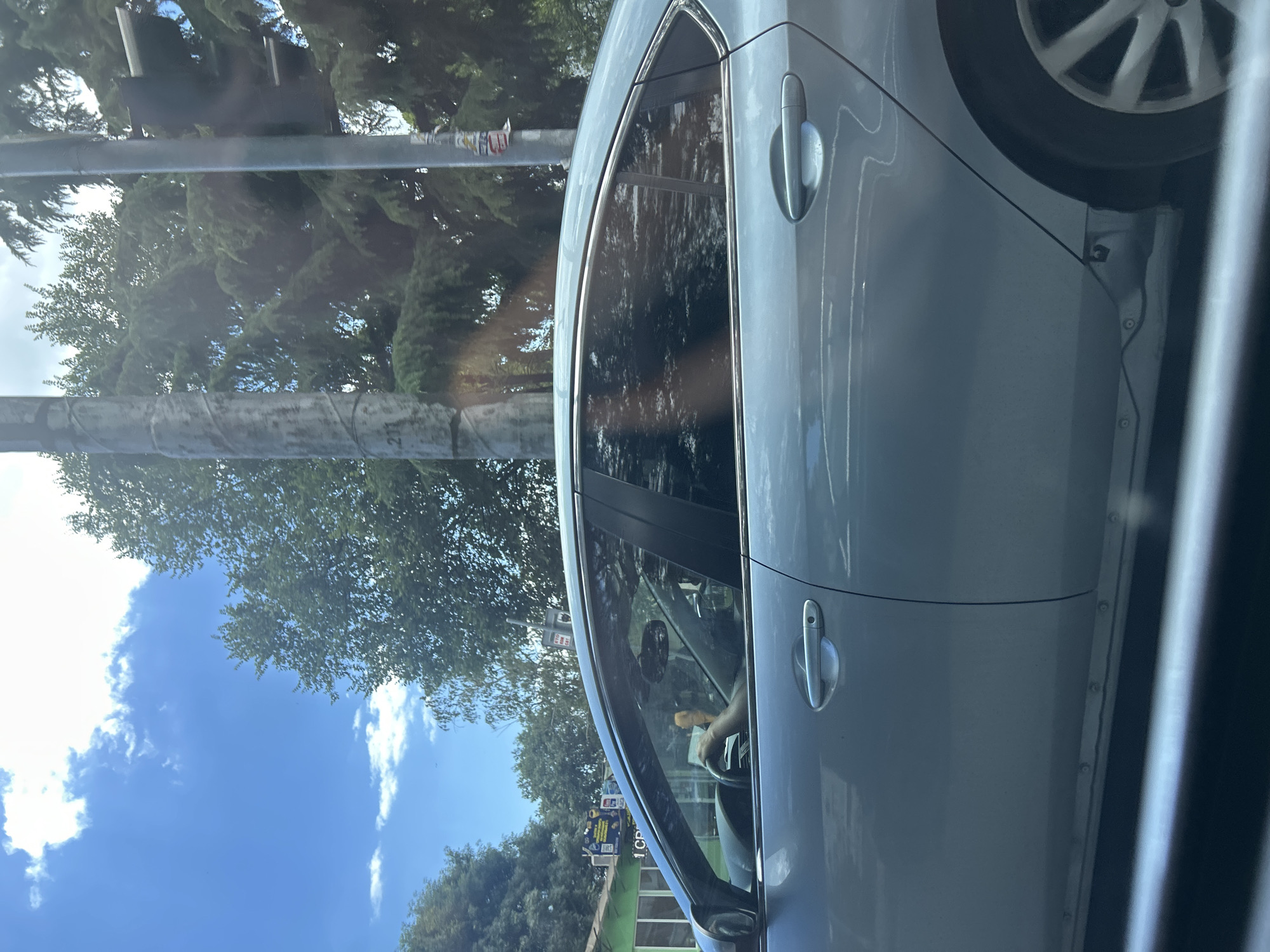 The image shows the side profile of a light blue sedan driving on a street in Varna, Bulgaria. The driver, with an arm resting on the steering wheel, is visible through the tinted windows. Trees and a traffic light are visible in the background, along with a blue sky with scattered clouds. The car appears to be moving, as suggested by the slight blur and the context of it being on a street. The environment suggests a sunny day in an urban or suburban setting.