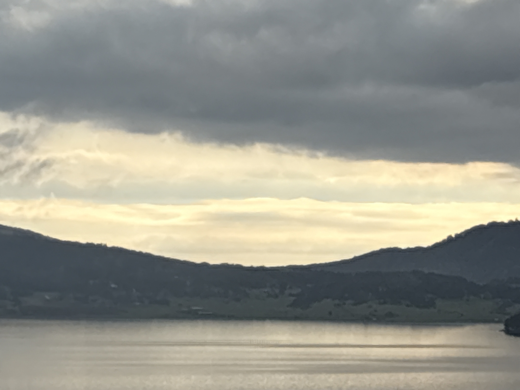 The image shows a landscape view of Tsigov Chark, Bulgaria. In the foreground is a large body of water, its surface slightly rippled with horizontal lines reflecting the light. Beyond the water, two landmasses or hills are visible, connected to create a valley shape. The hills are covered with trees. The sky is overcast with gray clouds. In the middle of the sky, there is a horizontal strip of brighter light.