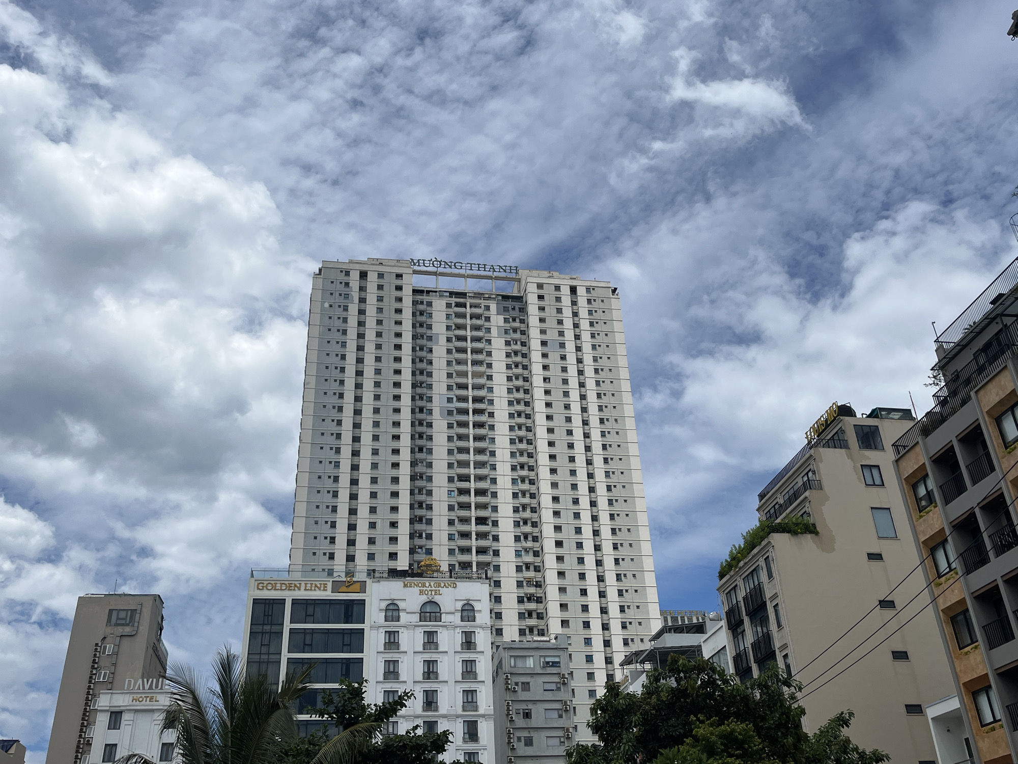 The image displays an urban landscape in Đà Nẵng, Vietnam, characterized by multiple multi-story buildings under a partially cloudy sky. The central and most prominent feature is a tall, slender high-rise building, light grey and white in coloration, featuring numerous uniformly arranged square windows and balconies. The name "MƯỜNG THANH" is affixed in blue capital letters to the rooftop structure of this building.
To the left of the central MƯỜNG THANH building, a white building with "GOLDEN LINE" in gold lettering on its upper facade is visible, featuring dark-framed windows. Further left, a taller, muted brown building with "DAVID HOTEL" in white lettering below its roofline is partially observed.
To the right of the MƯỜNG THANH tower, a white building with the inscription "MENORA GRAND HOTEL" in gold letters is present, distinguished by arched windows on its upper floors and balconies. Adjacent to it, a grey building with multiple external air conditioning units is seen. Further to the right, a light brown building with black-railed balconies and some rooftop vegetation is visible, alongside a segment of another building with a darker brown facade.
Green foliage, including palm fronds, is situated in the lower foreground, partially obscuring the bases of some structures. Black electrical wires are visible spanning horizontally across the mid-right section of the image. The sky above features scattered white cumulus clouds against a blue background. No individuals are present in the scene.