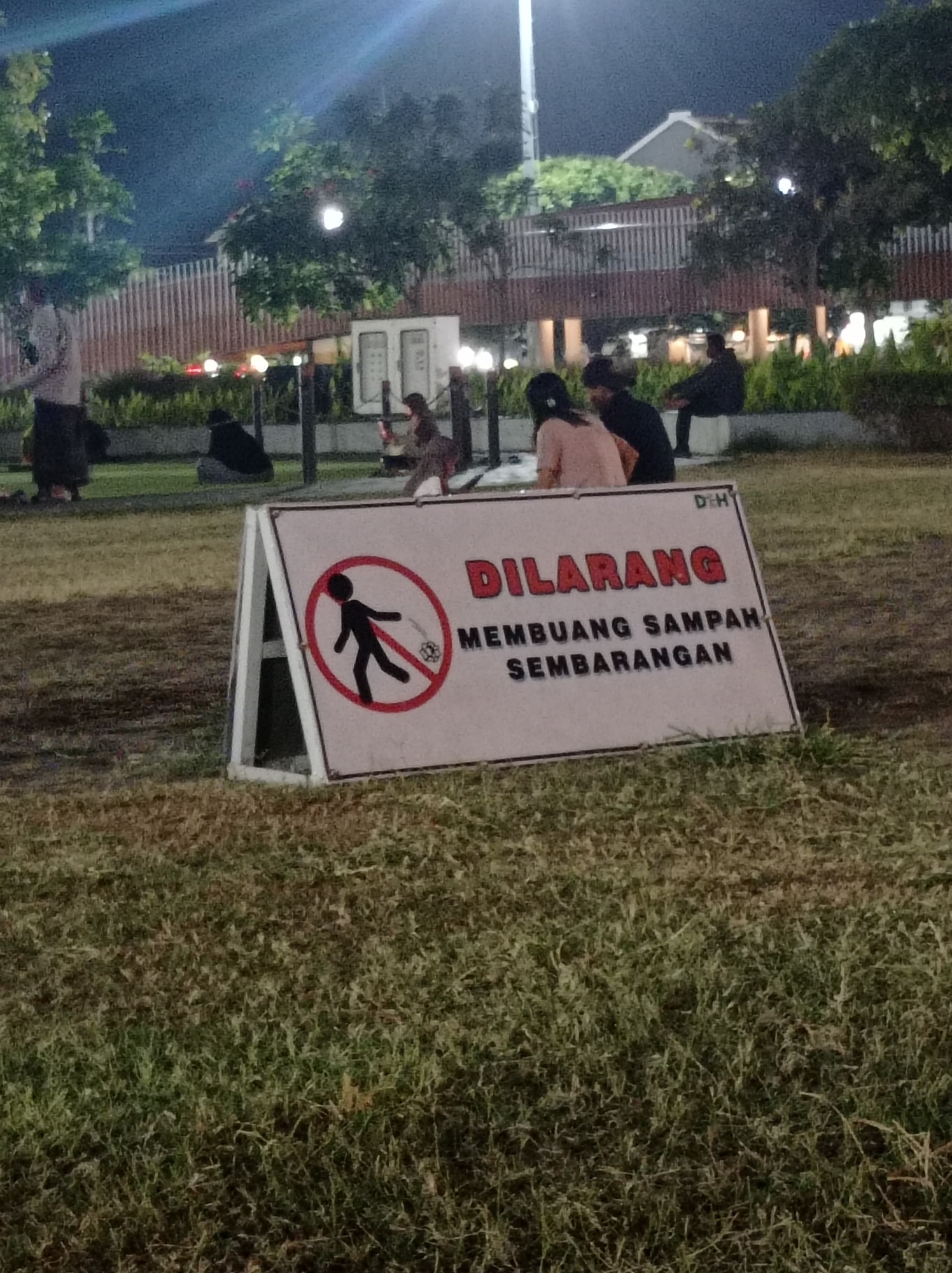 The image depicts a public park area at night in Tegal, Indonesia. In the foreground, a white A-frame sign displays a prohibition symbol with a silhouette of a person throwing trash. Text on the sign reads "DILARANG MEMBUANG SAMPAH SEMBARANGAN," which translates to "DO NOT LITTER." The park features a grassy lawn, trees, and various structures in the background, including lighting and what appears to be a covered seating area. Several individuals are visible in the middle ground, some seated on benches or the ground, while others are standing. The overall atmosphere suggests a recreational space at dusk or after dark, illuminated by artificial lighting.