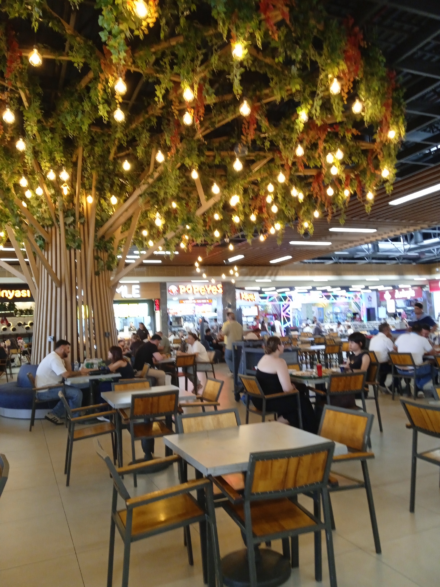 The image depicts an indoor food court area in Yenişehir, Turkey, characterized by a large, artificial tree structure adorned with numerous Edison-style light bulbs and faux greenery. The food court features multiple tables and chairs arranged throughout the space, with several people seated and dining. In the background, various food establishments are visible, including Popeyes. The overall atmosphere appears casual and bustling.