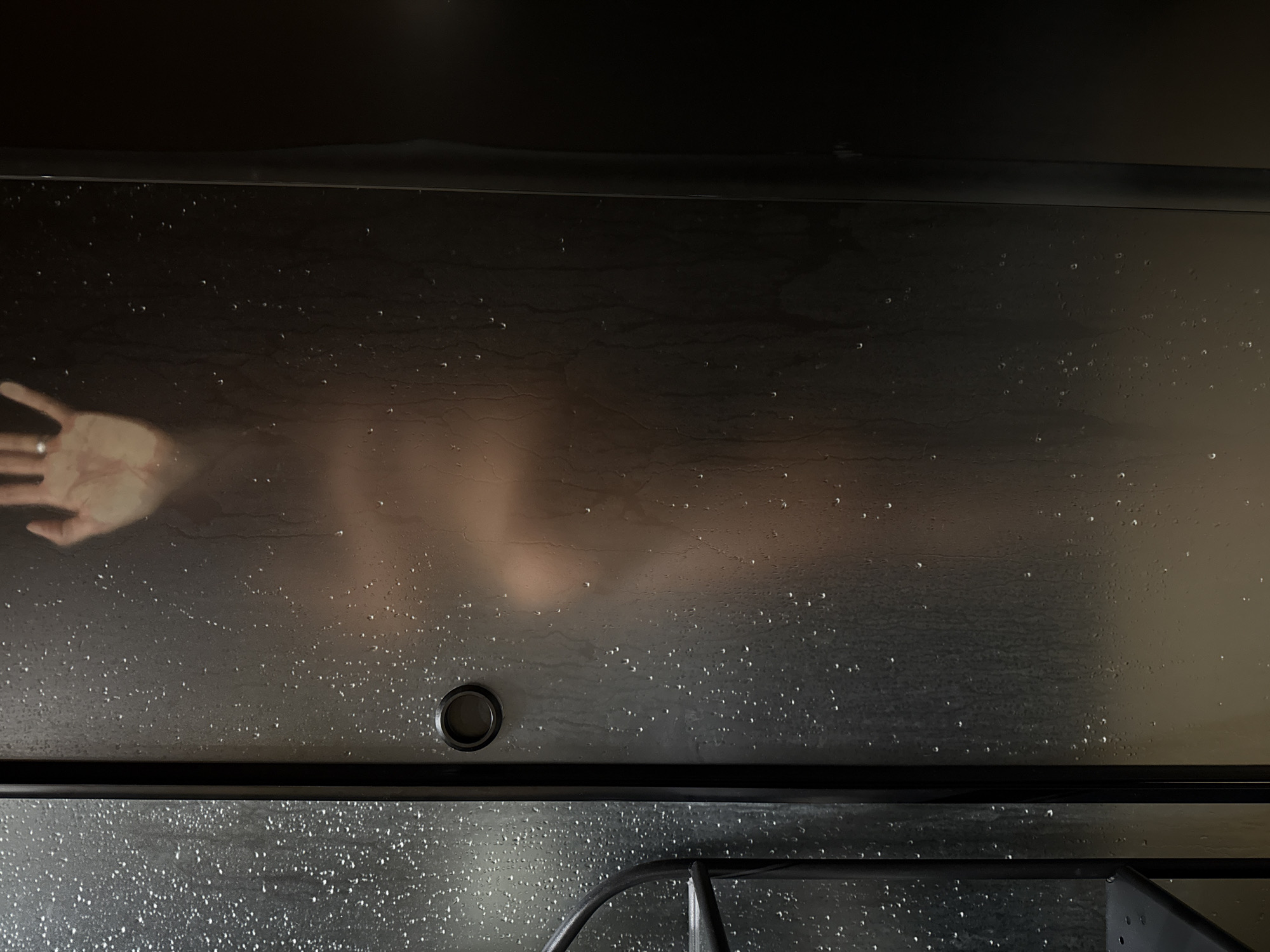 The image shows a person's torso and hand pressed against a frosted glass shower door. Water droplets are visible on the glass. The person's body is partially obscured by the frosted glass, but appears to be unclothed. The hand is raised and touching the glass, with a ring visible on one finger. The lighting is dim, creating a shadowy and intimate atmosphere. The image does not provide any information about a location in Chaikovsky, Russia.