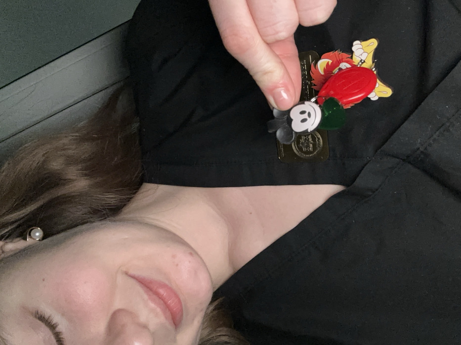 A woman is pictured, smiling slightly with light skin and brown hair. She is wearing a black uniform, possibly scrubs. Held above her uniform is a hand pinching a cluster of Disney-themed pins. The pins include Mickey Mouse, a red heart, a green shamrock, and a character resembling Mufasa from The Lion King. The photograph is taken at an angle, suggesting she is lying down while capturing the image. An earring with a pearl is visible on her left ear.