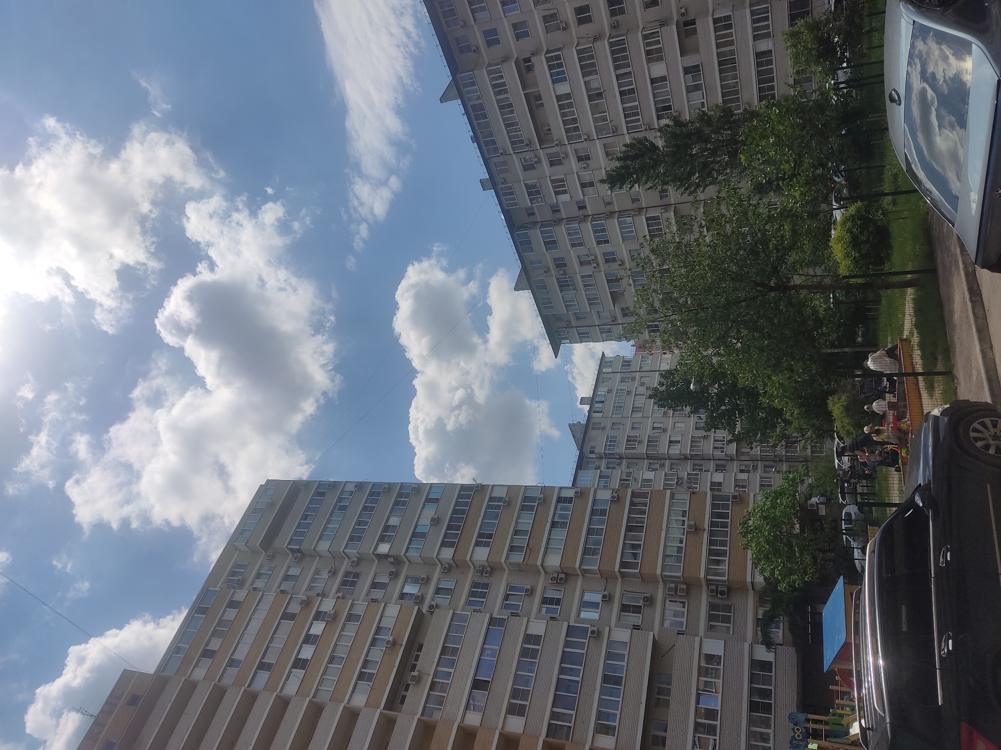 The image shows an upward, angled view of apartment buildings against a blue sky with scattered clouds. The buildings are tall and have a repetitive pattern of windows and balconies. Some trees are visible between the buildings. Cars are parked on the street and a playground can be seen. People are sitting on benches. The photo was taken in Krasnodar, Russia.