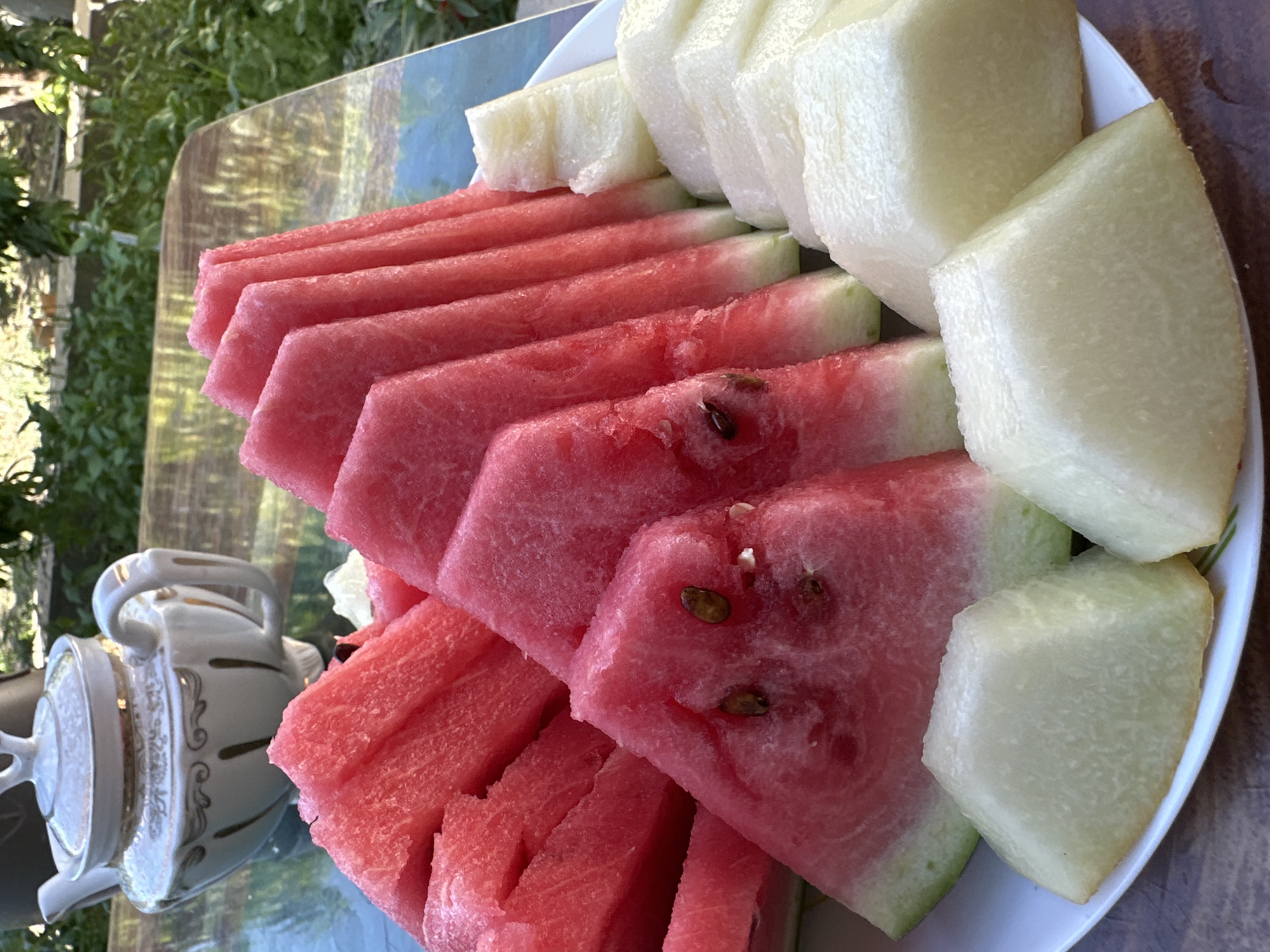 A plate of sliced watermelon and honeydew melon is arranged on a table. The watermelon slices are a vibrant pink, with visible seeds. The honeydew melon slices are pale green and appear firm. To the left of the plate is a white teapot with gold trim, sitting on a reflective surface that appears to be a glass-topped table. In the background, lush green foliage suggests an outdoor setting. The overall scene is bright, implying daytime. There is no visible text in the image.