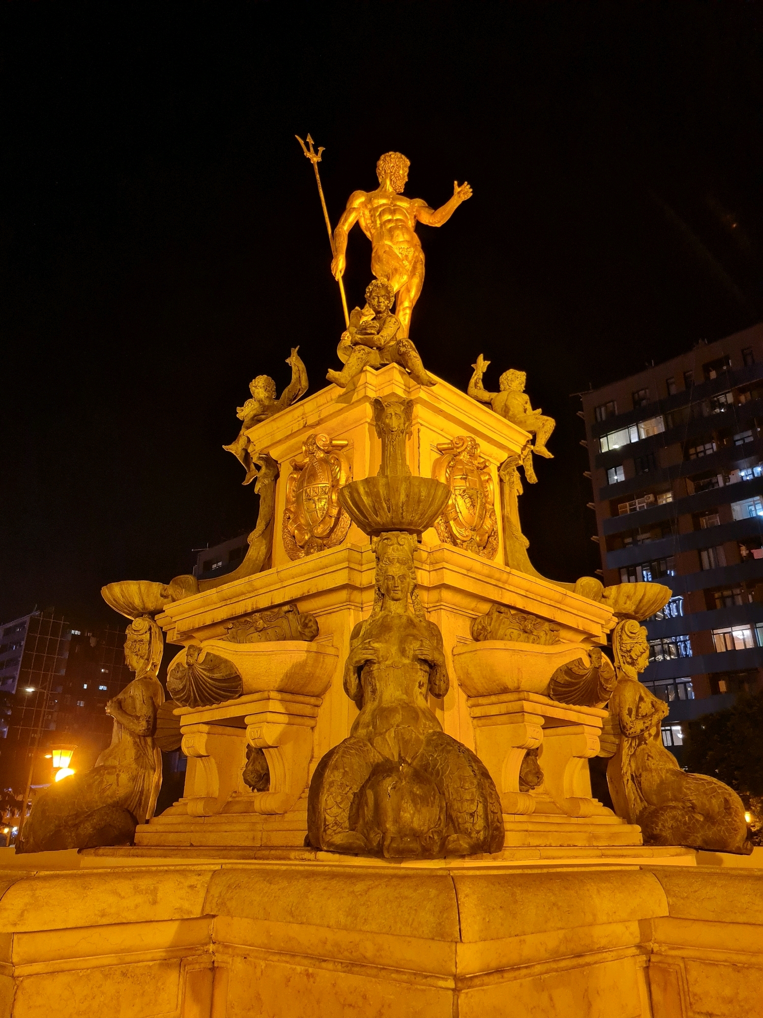 The image depicts the Neptune Fountain in Batumi, Georgia, at night. The central subject is a golden statue of Neptune, the Roman god of the sea, standing atop a multi-tiered fountain. Neptune holds a trident in his right hand and gestures with his left. Surrounding him are several smaller sculptures, including cherubs and figures with tails, possibly representing mythical sea creatures or sirens. The fountain itself is ornately decorated with reliefs and architectural elements. The scene is illuminated by artificial lighting, casting a warm glow on the golden statue and the stone structure. In the background, a tall apartment building with illuminated windows is visible, along with streetlights and some trees. The overall atmosphere is one of grandeur and historical significance, set against the backdrop of a modern urban environment.