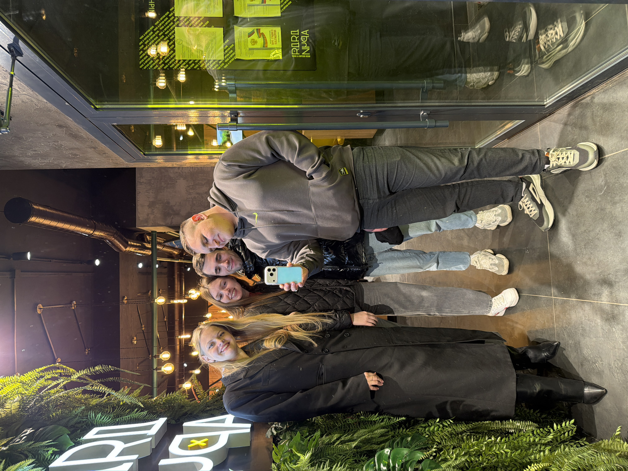 A group of four people is posing for a selfie in front of a business. A young blonde woman in a black coat stands closest to the camera, next to a sign with green foliage. Behind her are two women, one holding a turquoise phone, and a man in a gray hooded sweatshirt. The building has a glass front. The sign behind the group says "RARI PUGA VEGAN" in white lettering. The location is likely Izhevsk, Russia, based on Cyrillic script on the window. The group is smiling and looking at the phone. The light fixtures can be seen inside the building. The floor appears to be made of tiles.