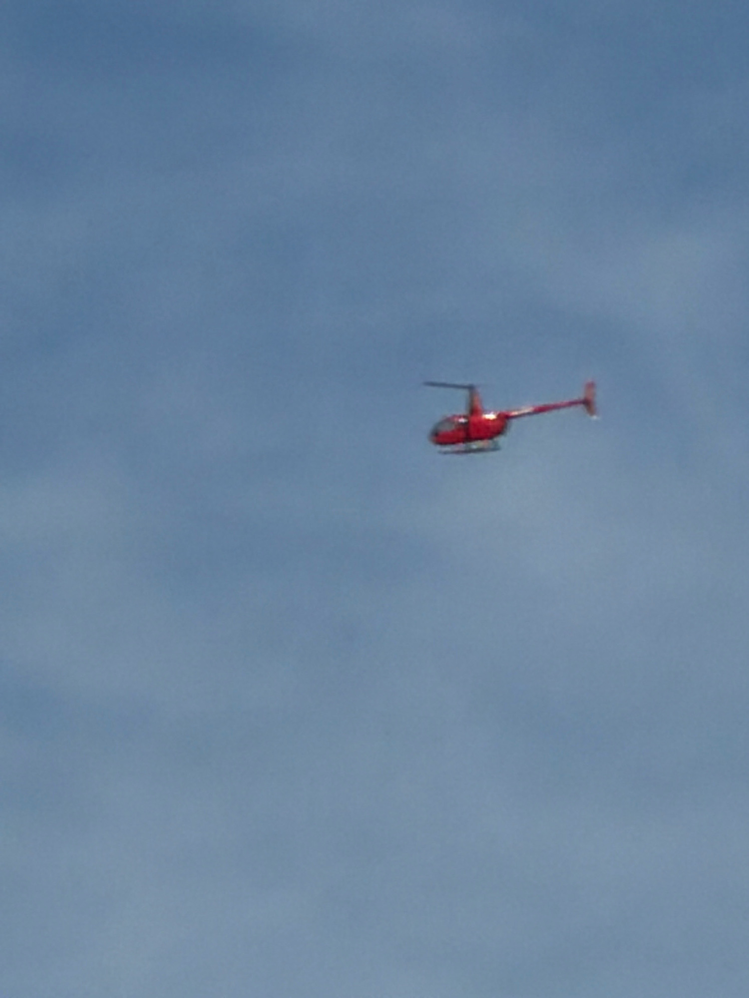 A red helicopter is flying in a clear blue sky. The helicopter is the only object visible in the image. The helicopter is small and appears to be a model or a remote-controlled helicopter. There are no people or buildings visible in the image. The image is taken from a low angle, looking up at the helicopter.