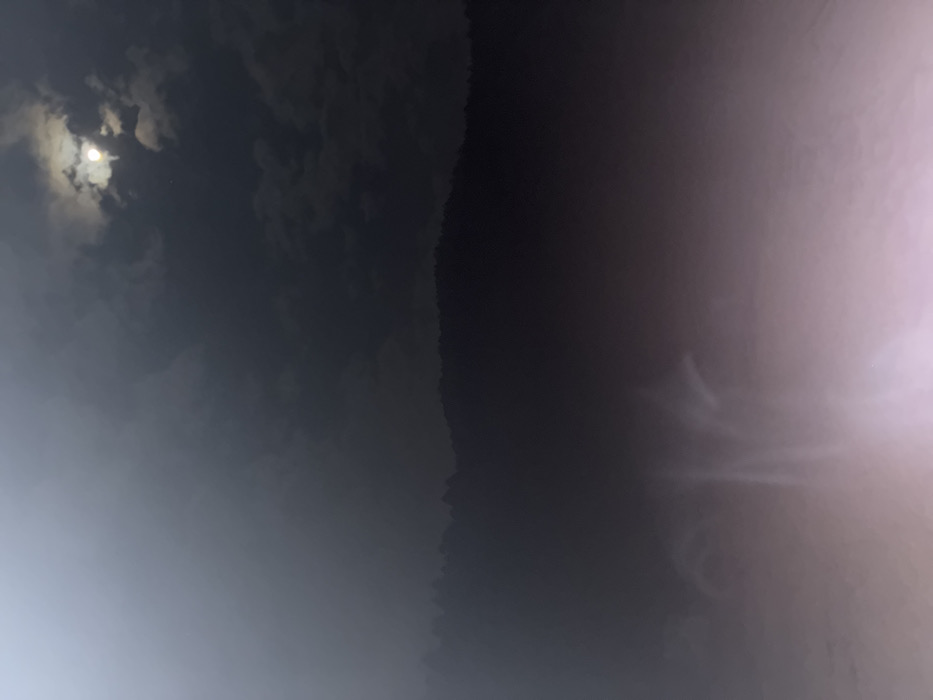 The image depicts an outdoor, low-light environment at night or under heavily overcast conditions in La Marquesa, Mexico. The upper portion of the frame shows a dark sky with significant cloud cover. A bright, diffused light source, consistent with a moon, is partially visible through gaps in the clouds in the upper right quadrant. Below the sky, a dark, silhouetted horizon line of what appears to be a forested mountain range or dense tree line extends across the midground. The lower half of the image is dominated by a diffuse, light-emitting haze, likely fog or ground-level mist. Within this haze, several elongated, upward-tapering luminous streaks are visible, resembling plumes of light reflecting through atmospheric conditions. There are no discernible people, specific objects beyond the natural landscape and atmospheric phenomena, or explicit actions or interactions depicted. The color palette transitions from dark blues and greys in the sky to pinkish-purple and white in the hazy foreground.