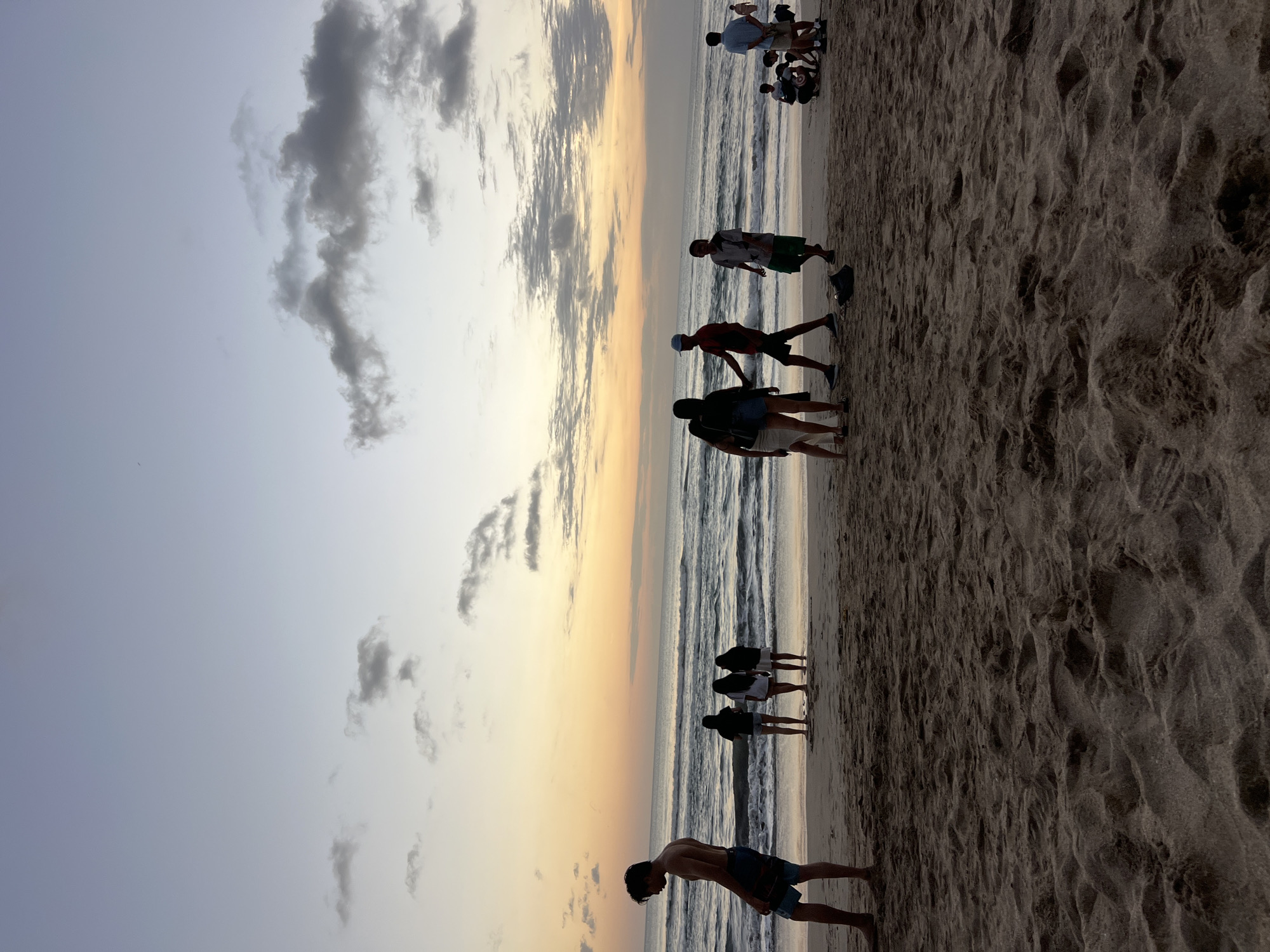The image captures a beach scene in Kuta, Indonesia, during what appears to be sunset or sunrise. The sky displays a gradient of colors, from a pale blue at the top to warm oranges and yellows near the horizon. Clouds are scattered across the sky, some appearing dark and textured.
The foreground is dominated by the sandy beach, showing footprints and the texture of the sand. In the middle ground, several groups of people are visible on the beach. On the left, a shirtless young man in blue swim trunks stands facing away from the camera, looking towards the water. Further along the beach, a group of three women stand together, their backs to the viewer. To their right, a group of three young men are interacting. One, wearing a red shirt and a cap, appears to be talking to another in a gray t-shirt and green shorts. Another person in a blue shirt stands further to the right. In the far right of the image, a small group of people are gathered, some appearing to be seated.
The ocean is visible in the background, with gentle waves rolling towards the shore. The overall atmosphere of the image is tranquil and suggests a casual gathering of people enjoying the beach at dusk or dawn.