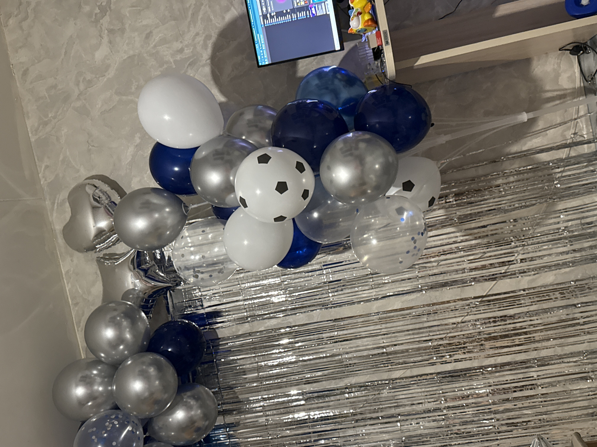 The image shows a balloon arrangement with silver fringe in an indoor setting, likely in Chaikovsky, Russia. Balloons are predominantly silver, white, and dark blue. One white balloon has black soccer ball markings. The arrangement is positioned against a wall with a textured, mottled gray pattern. A television screen and a shelf are visible in the upper portion of the image. The overall lighting is somewhat dim.