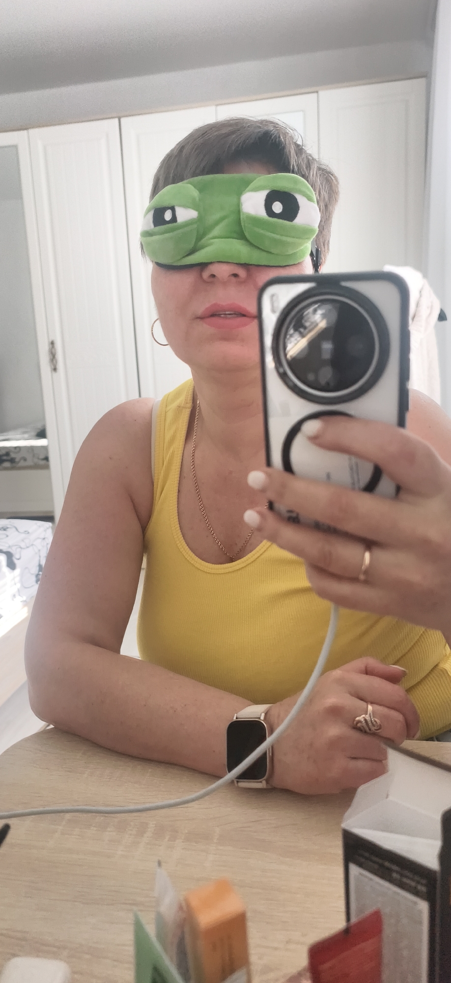 A woman is taking a selfie with a smartphone while wearing a green frog eye mask. She is wearing a yellow tank top, a gold necklace, a gold ring on her right hand, and a smartwatch on her left wrist. The location appears to be indoors, with a white closet in the background. The smartphone is connected to a white charging cable. Various boxes are visible on the table in front of the woman. There is no indication that the image was taken in the city of Фоки, Russia.