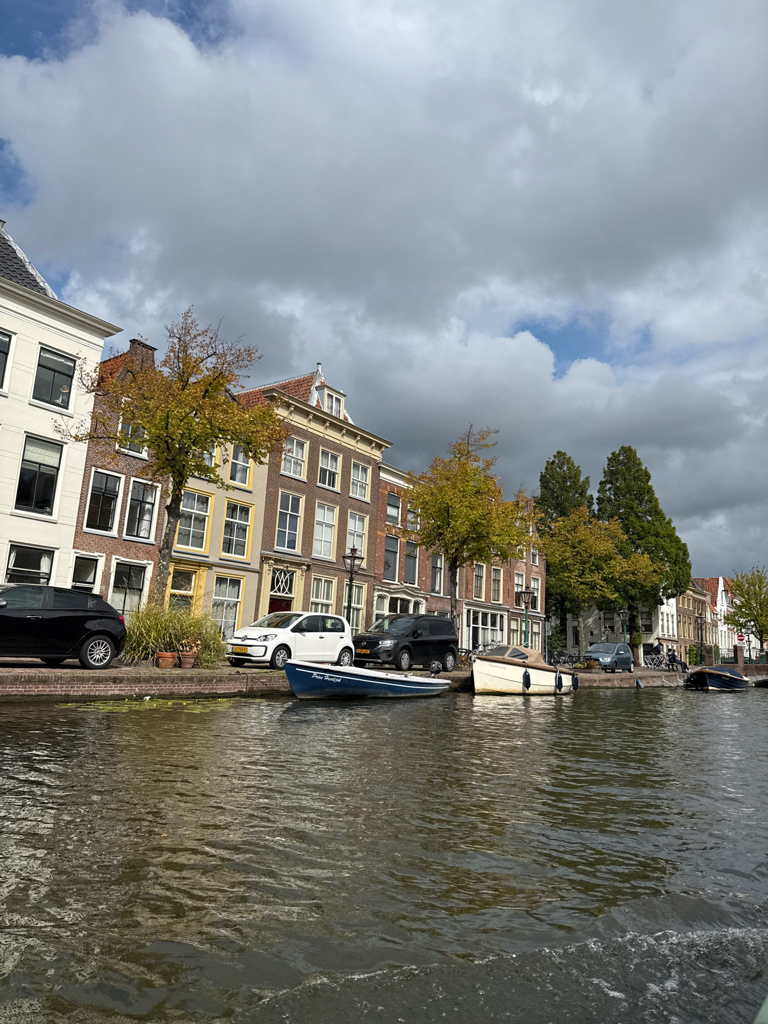 This image captures a canal-side view of historic buildings in Leiden, Netherlands, likely along the Rapenburg canal, given the architecture and canal type. The buildings are a mix of styles, with brick facades, large windows, and pitched roofs, indicative of Dutch Golden Age architecture. Several cars are parked along the canal bank, including a black hatchback, a white hatchback, and a darker colored SUV. A few boats are moored in the canal, one a small blue vessel with text that appears to read "Prins Hendrik". Another is a larger, cream-colored boat with a covered section. Trees with yellowing autumn leaves are visible along the canal, suggesting the season is fall. The sky is overcast with dramatic clouds, with a few patches of blue sky peeking through. The canal water is murky and shows ripples, suggesting movement. There are a few people visible in the distance, some sitting on benches and others walking along the canal path.