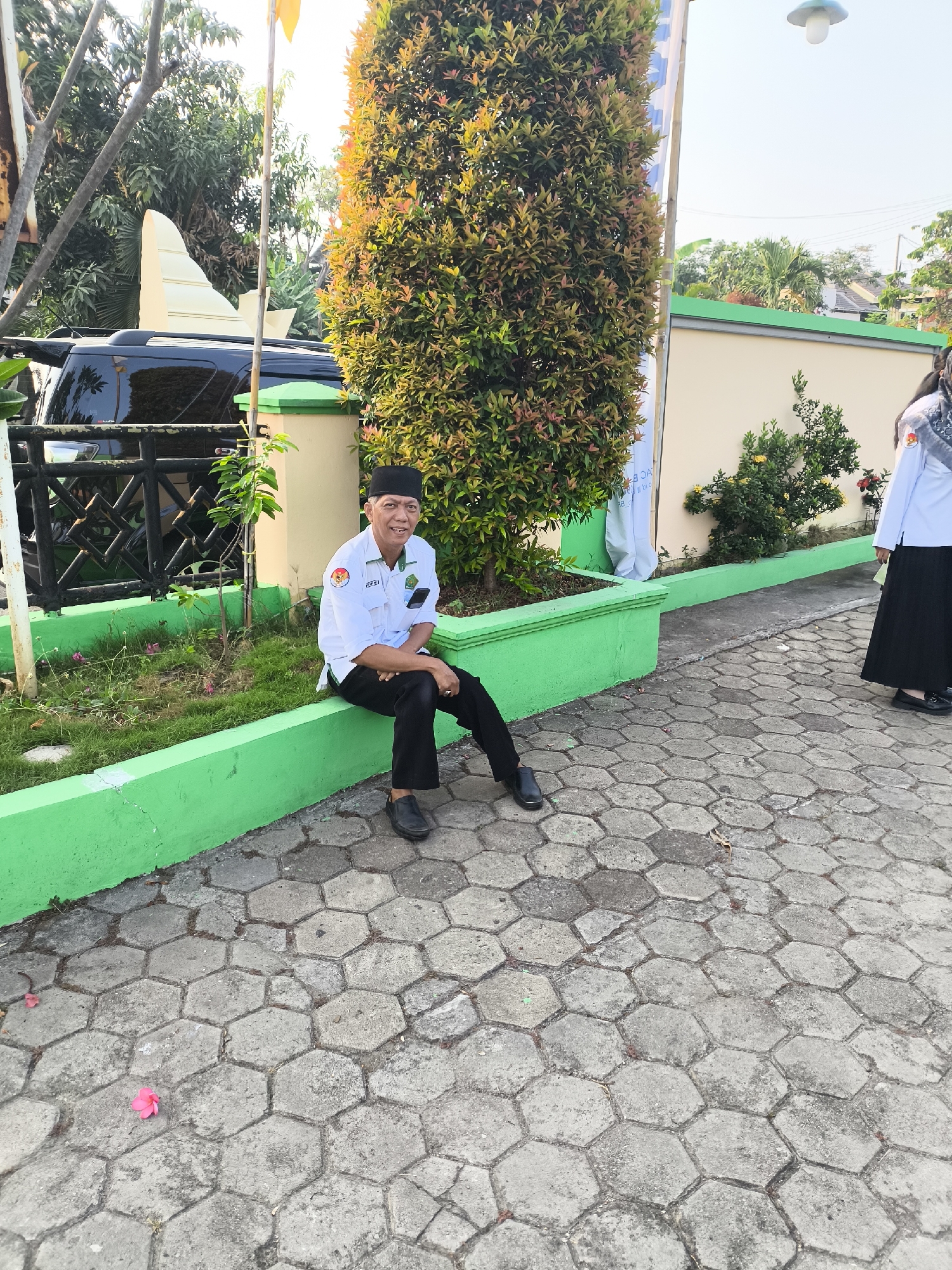An outdoor scene in Cilegon, Indonesia features a man in a white uniform and black pants sitting on a bright green concrete border. He wears a black cap and black shoes. Behind him, a bushy tree with red and green foliage stands tall. A black SUV is parked behind a black metal fence to the left of the man. A light-colored building with a green trim runs along the background, with trees and other vegetation visible. The ground is paved with hexagonal grey concrete blocks, with a few fallen pink flowers scattered on the ground. A portion of another person in a white uniform and black skirt is visible to the right.
