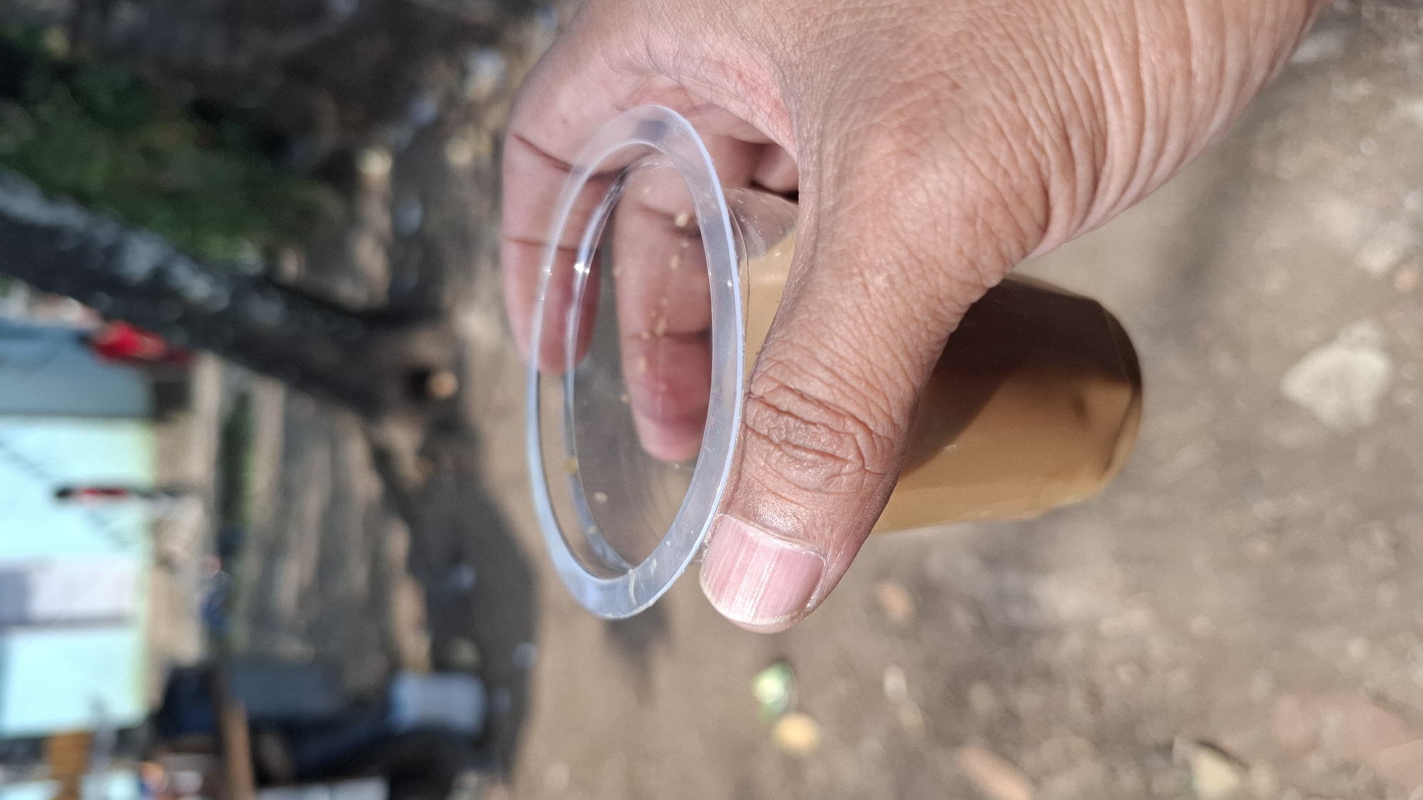 In the foreground, a hand with visible fingernails and textured skin grips a clear plastic cup containing a light brown liquid, possibly tea or coffee. The cup has a clear plastic lid with a raised rim. The background is blurred, showing an outdoor environment. To the left, a wooden structure is partially visible, with a white bucket underneath. In the middle ground, a tree trunk is prominent. Further in the background, a building with a light blue wall and a red figure standing near it are discernible. The ground appears to be a mixture of dirt and scattered debris. The scene is likely in a public or street setting within the city of Cilaku, Indonesia, given the architectural style and general atmosphere.