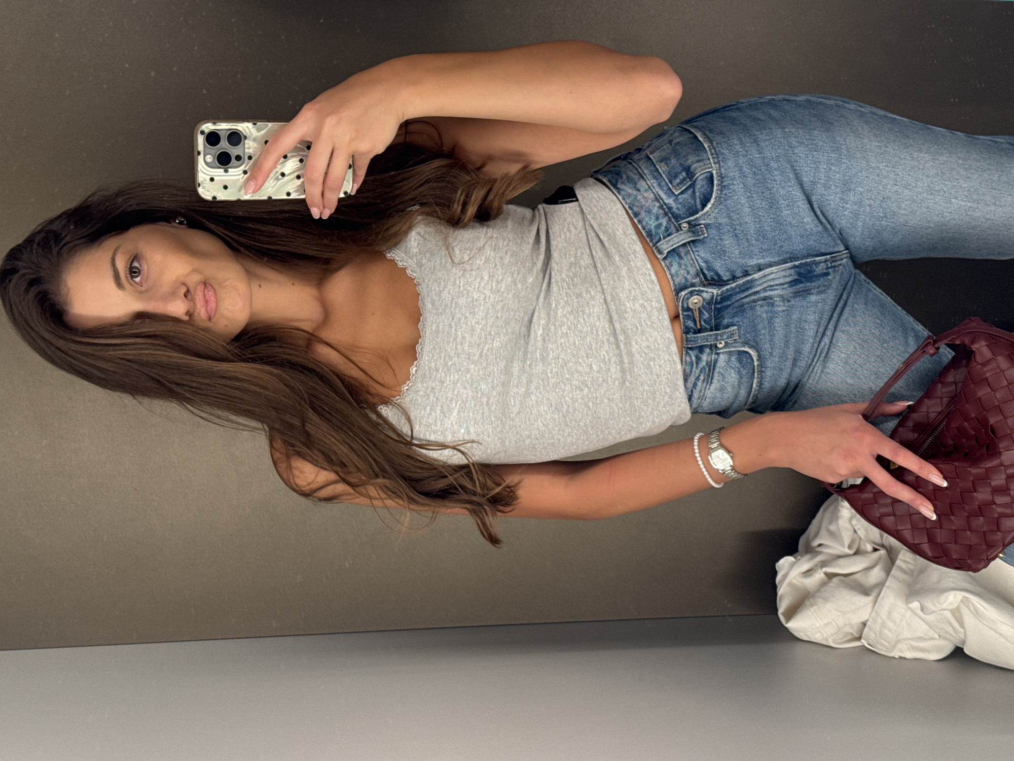 A young woman takes a selfie indoors. She is wearing light blue jeans and a gray top with lace trim. She holds a modern smartphone in her left hand and a burgundy purse in her right hand. There is a pile of clothing to her right. The background consists of a gray wall. It is not possible to identify the location in Burgas, Bulgaria from the image.