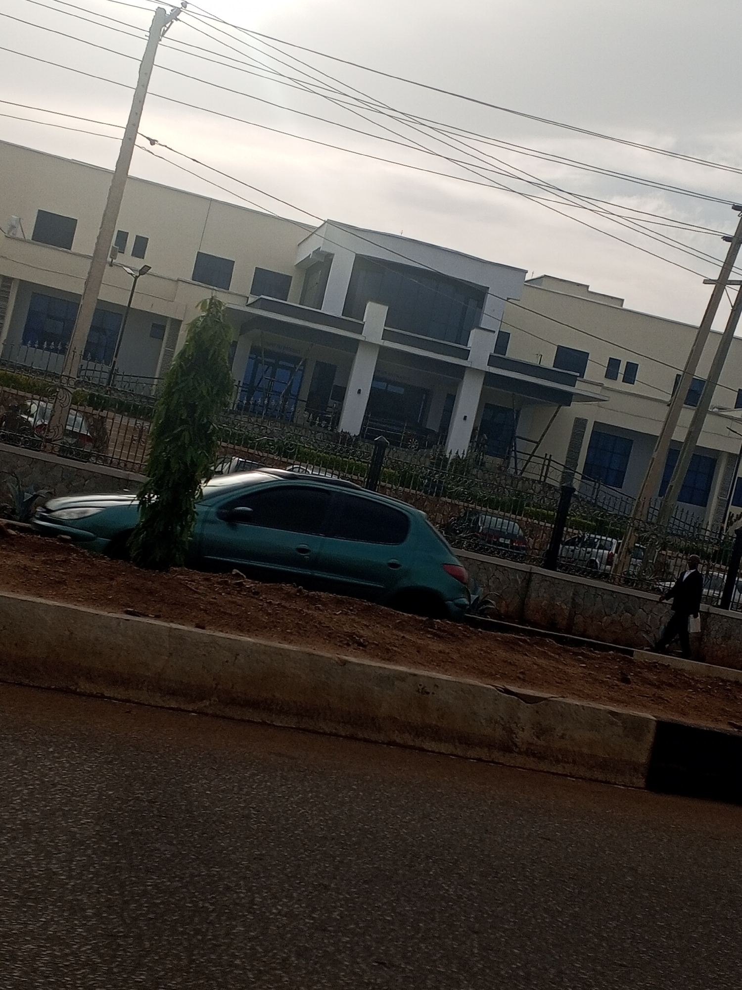 A teal colored car is partially submerged in dirt, with its front bumper resting on the ground. The car is situated on an incline, with a thin, tall evergreen tree growing to its left. Behind the car, a black metal fence encloses a property with a modern building. The building has a light-colored facade with dark tinted windows and a prominent entryway. Power lines are visible crisscrossing the sky above. A single person in a dark suit walks on the sidewalk to the right of the fence. The sky is overcast, suggesting a cloudy day. The scene appears to be an urban or semi-urban setting, possibly in Jos, Nigeria, given the context. The overall mood is somewhat bleak due to the car's predicament and the overcast weather.