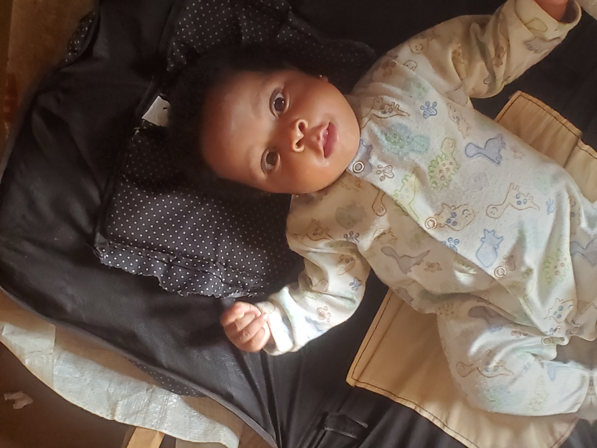 A baby lies on a dark gray surface with a black and white polka dot fabric nearby. The baby is wearing a patterned onesie with light-colored animal designs. A tan fabric is also partially visible. The location and the city of Ukwu-Abwa, Nigeria are unable to be determined by visual analysis.