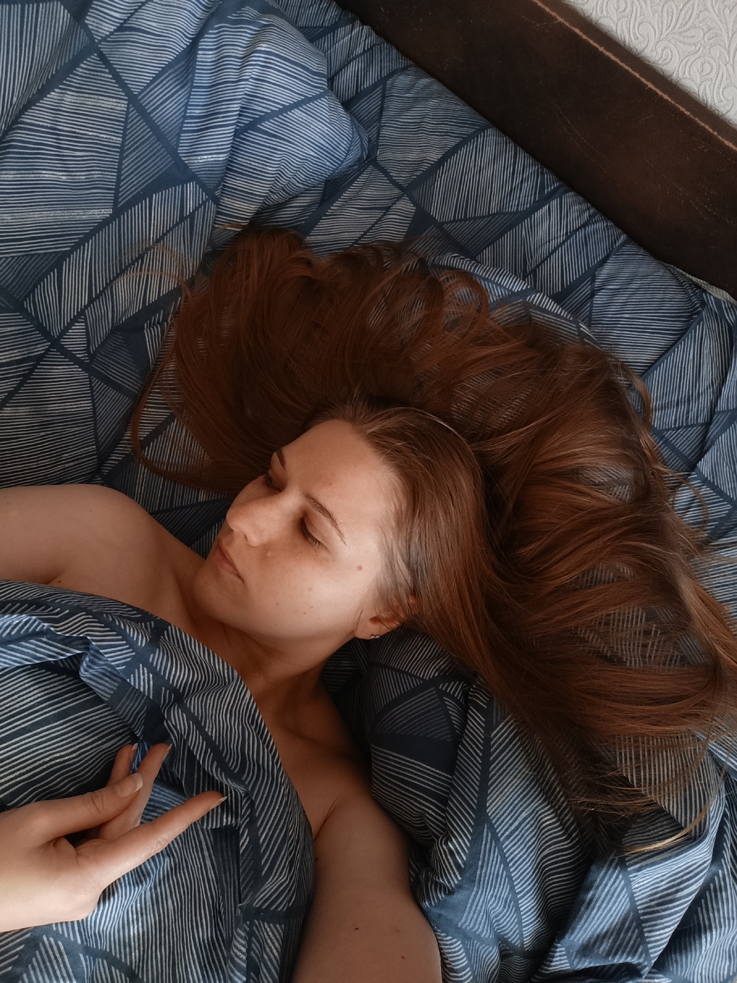 A woman with long auburn hair is lying in bed on her side, eyes closed and her head resting on a pillow. She is covered by a blue duvet with a geometric pattern. Her left arm is visible, with her hand resting on the duvet. The scene appears to be indoors, possibly in a bedroom.