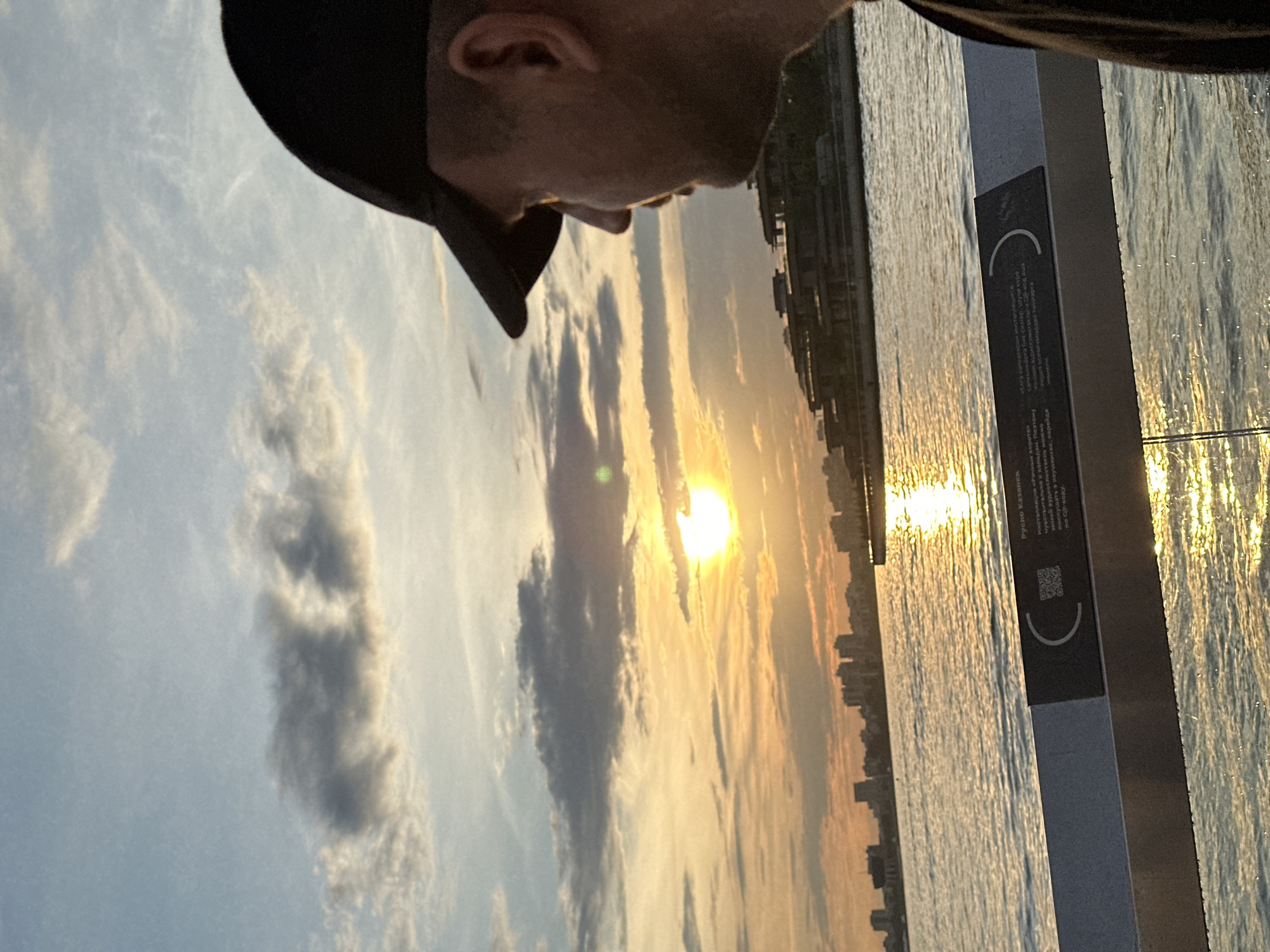 The image shows a man wearing a black baseball cap, seen in profile from the right, looking out at a sunset over the Kazanka River in Kazan, Russia. The sun is low in the sky, casting a warm golden glow on the water and clouds. In the background, a city skyline with modern buildings is visible, along with a distinctive building complex across the river. In the foreground, a railing with a plaque containing Cyrillic text and a QR code is present. The overall scene captures a peaceful, reflective moment against a vibrant natural backdrop.