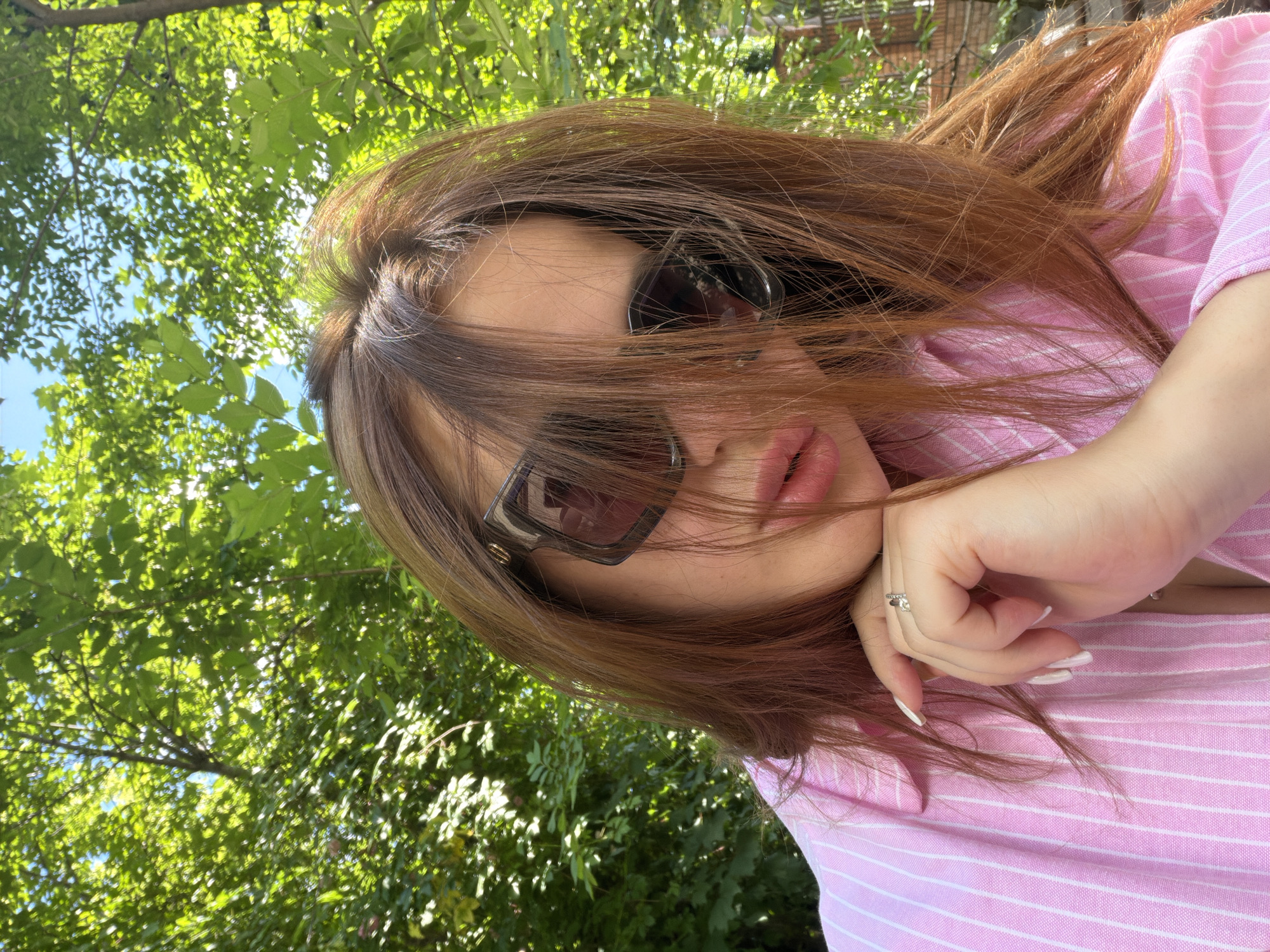 A young adult female is depicted in a medium close-up shot outdoors in Krasnodar, Russia. Her long, reddish-brown hair is parted and falls across her face, partially obscuring large, square-framed dark sunglasses. The left lens (viewer's right) of her sunglasses exhibits white marks. She wears a pink collared shirt with thin white vertical stripes. Her left hand (viewer's right), with white-painted nails and a ring featuring a clear stone on the ring finger, is positioned against her chin. Her lips are full and appear to have lip gloss applied. The background is dominated by bright green deciduous tree foliage under clear blue sky, indicating strong sunlight. A subtle reddish-brown brick structure is discernible in the far right background.