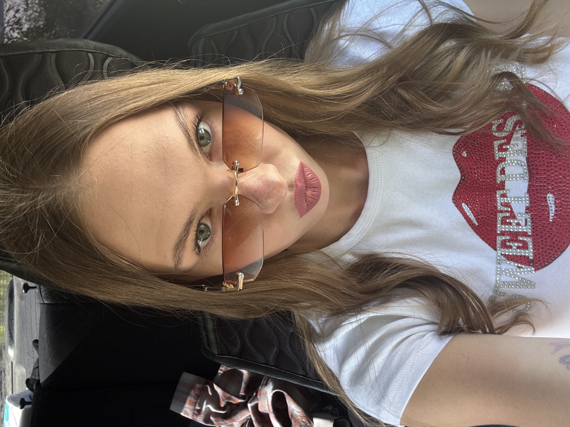 A young Caucasian woman with long, light brown hair is shown in a close-up, angled selfie, taken outdoors during the day. She is wearing a white t-shirt with a large, stylized red lip graphic embellished with red and silver rhinestones, and the word "SECRETS" is written in silver rhinestones across the lips. She is also wearing rimless sunglasses with brown lenses and gold frames. Her lips are painted a matte pink.
The setting appears to be inside a vehicle, with the back of a car seat visible in the upper left corner. The natural light suggests it is daytime. There are no obvious weather cues. The woman's expression is neutral, and the overall tone of the image is casual and self-expressive.