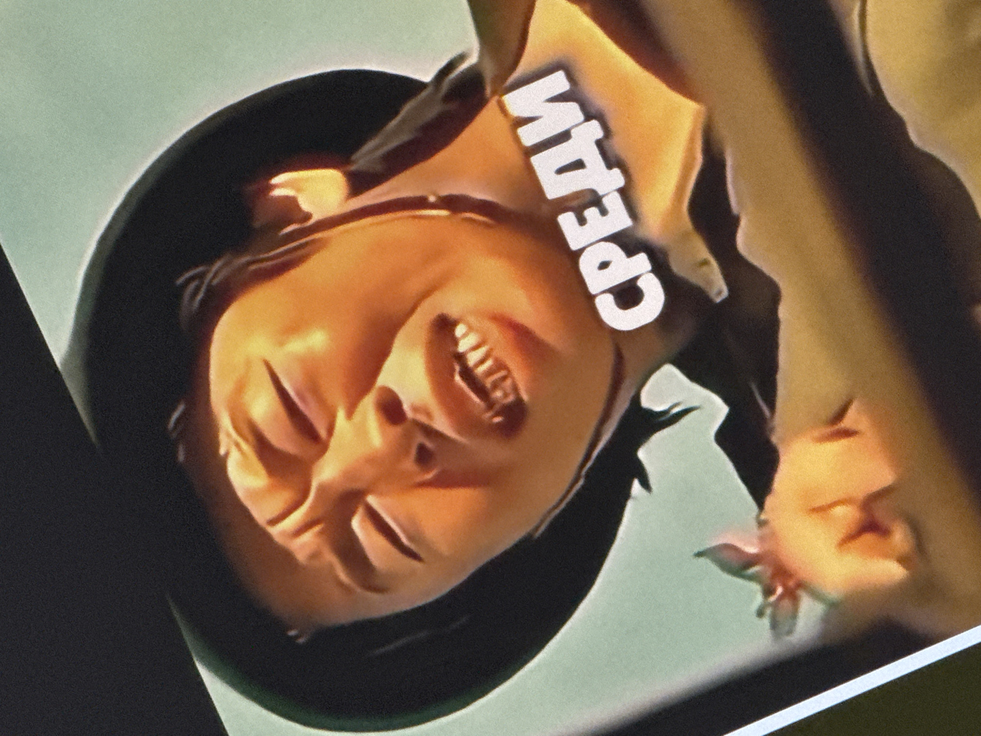 The image is a close-up of a person's face, wearing a dark hat. The person has their eyes closed and their mouth open, with teeth visible. There is text overlayed on the image that reads "СРЕДИ" in white, bold, 3D lettering. The background is a light blue-green color. The person appears to be an Asian male with dark hair. The lighting suggests the image might be from a screen or a photograph taken indoors or at dusk. There is no identifiable information to link this image to the city of Saratov, Russia.