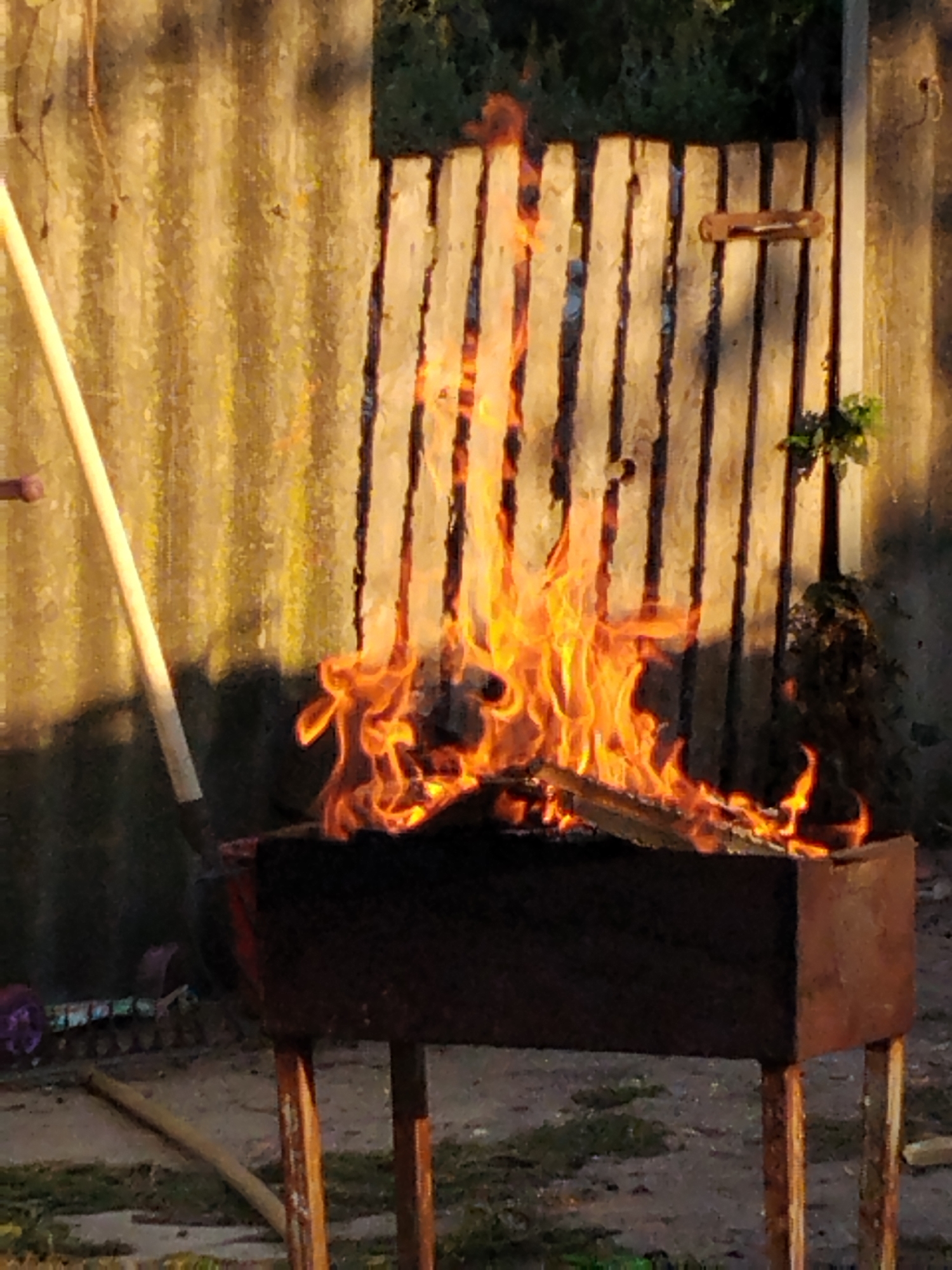 In Ustyvytsia, Ukraine, an outdoor scene features a fire burning in a dark, rectangular brazier supported by four wooden legs. The brazier, made of metal, is filled with logs that are engulfed in flames and smoke. To the left of the brazier, a long, light-colored pole, possibly part of a tool or equipment, leans against the flames. In the background, a rustic wooden fence with vertical planks stretches across the image. The fence is weathered, with some planks appearing darker than others, and a small, dark object resembling a bracket or holder is attached to one of the planks. Sunlight casts long shadows on the fence, suggesting it is either early morning or late afternoon. The ground around the brazier appears to be dirt and gravel. A glimpse of greenery can be seen behind the fence on the right side of the frame.